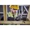 A construction worker in protective clothing, wearing a hard hat and safety glasses, is using a handsaw to cut wood. Scaffolding is visible in the background.