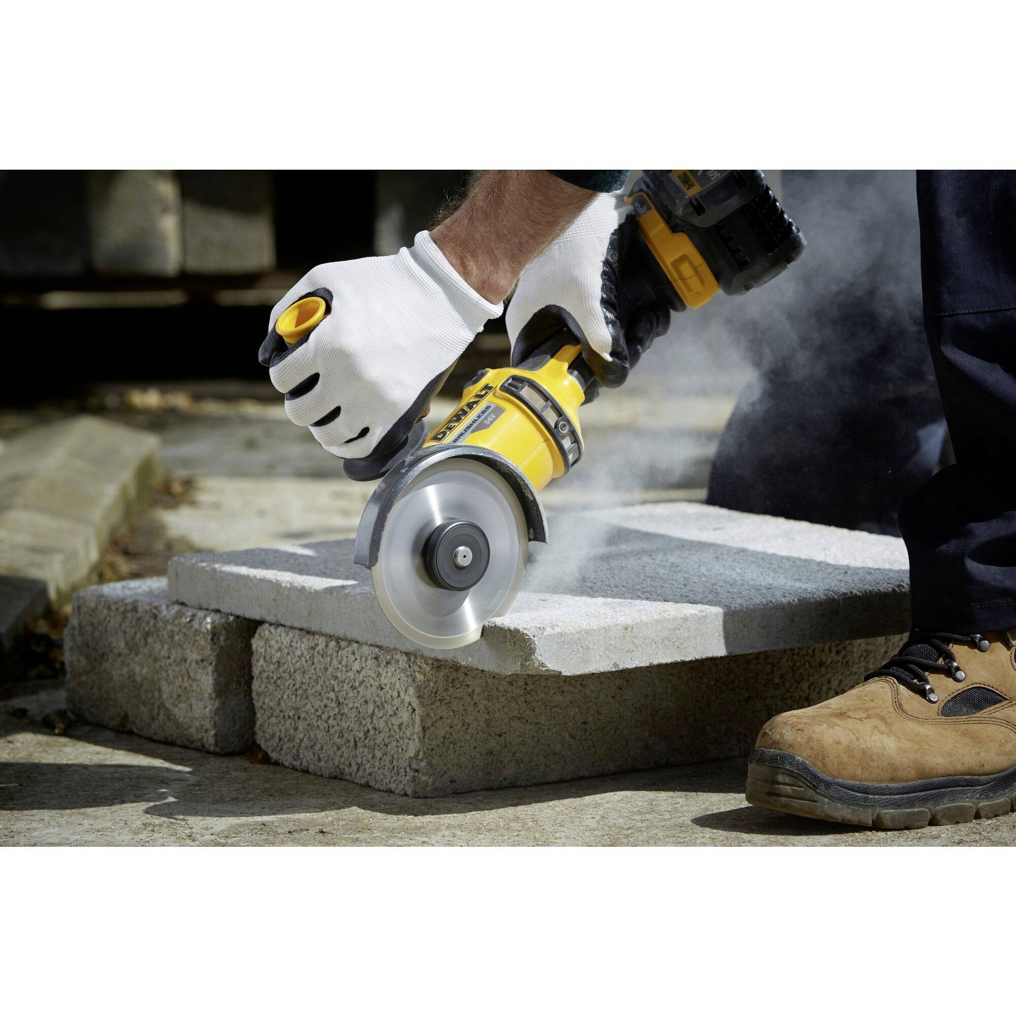 A worker is cutting concrete blocks with a handheld circular saw. Protective clothing and gloves are visible. Dust is being kicked up.