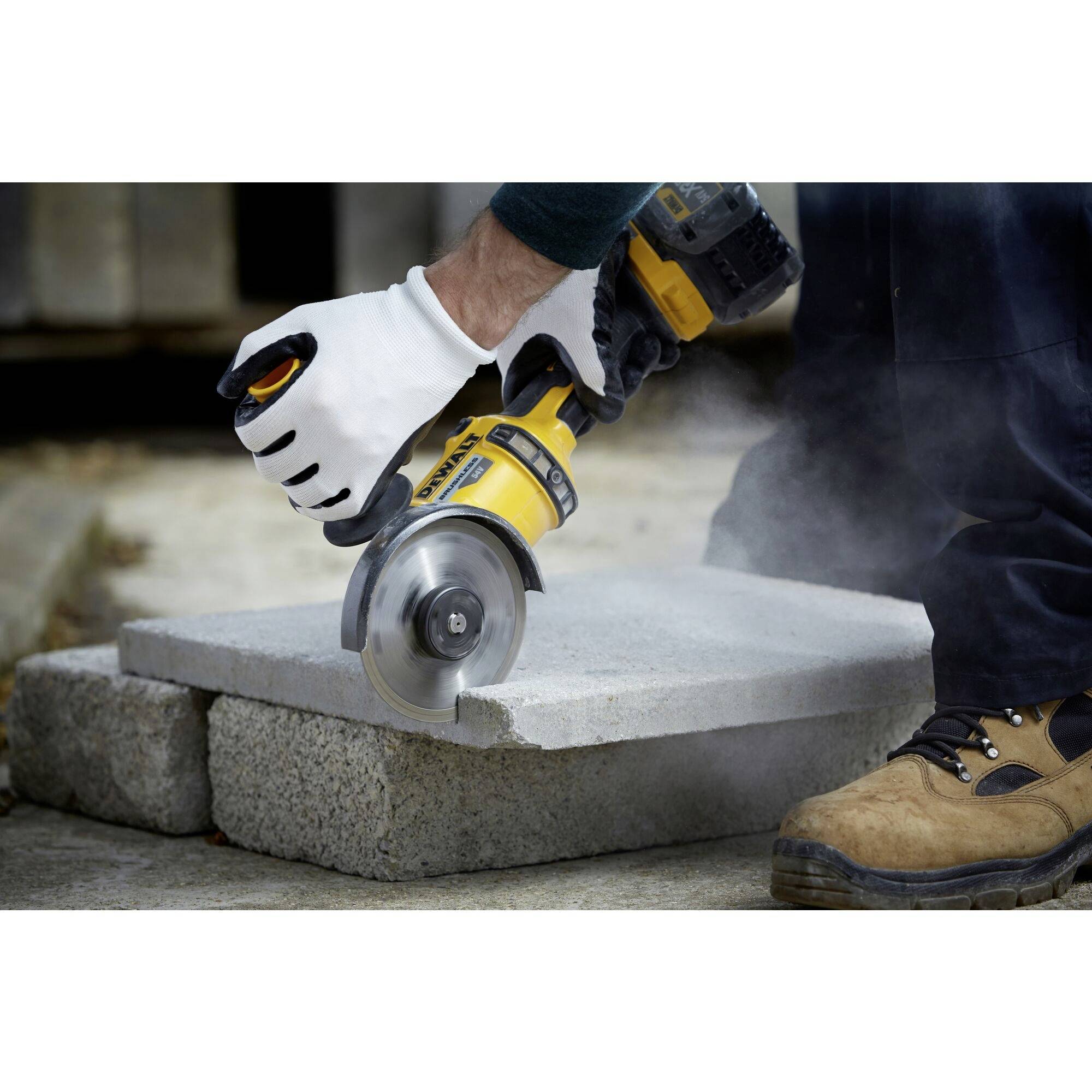 A person is cutting a concrete block with an electric saw. Dust clouds form during sawing. The person is wearing work gloves and boots.