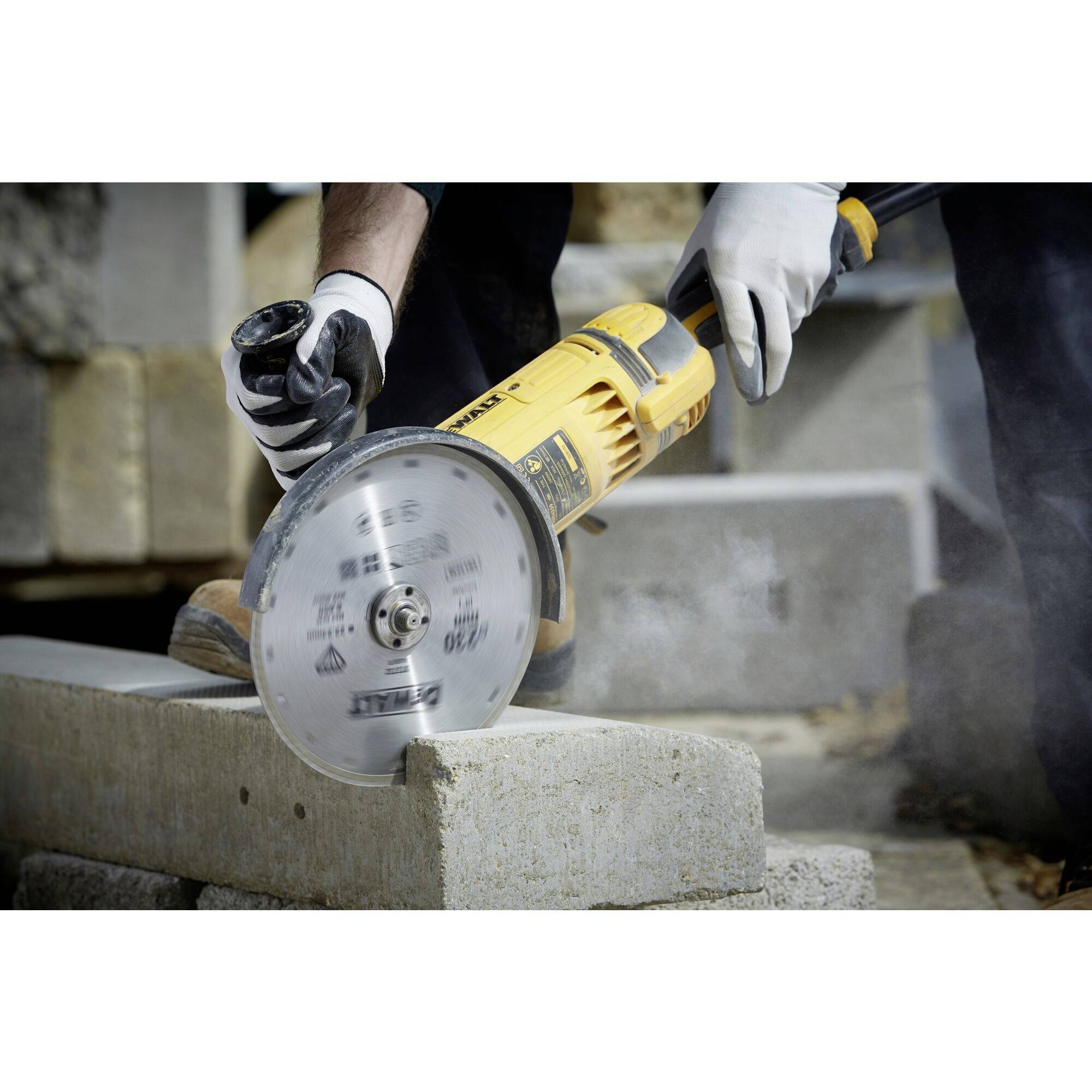A person is cutting concrete stone blocks with a yellow, electric cutting angle grinder. Dust is being generated in the process. Protective clothing is visible.