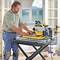 A man is using a table saw in a room with light-coloured walls and windows. Safety equipment and building materials are visible.