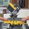 Two hands are operating a yellow mitre saw on a table. The saw is slightly angled. The background shows a kitchen.