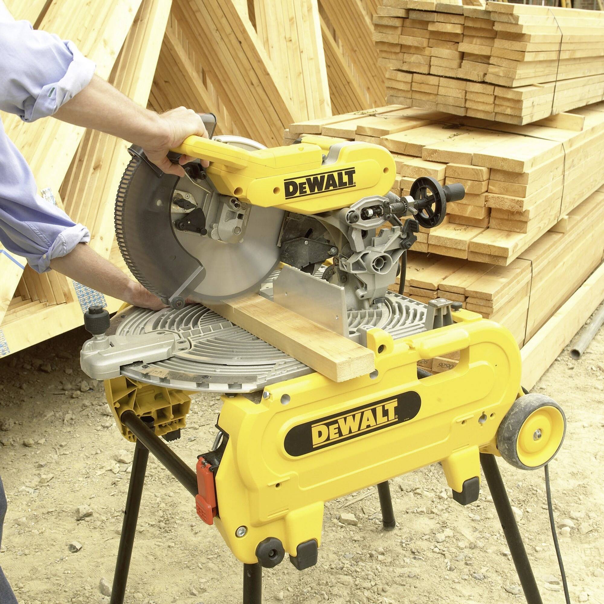 A person is sawing a wooden board with a yellow mitre saw on a building site. Stacked wooden boards are visible in the background.