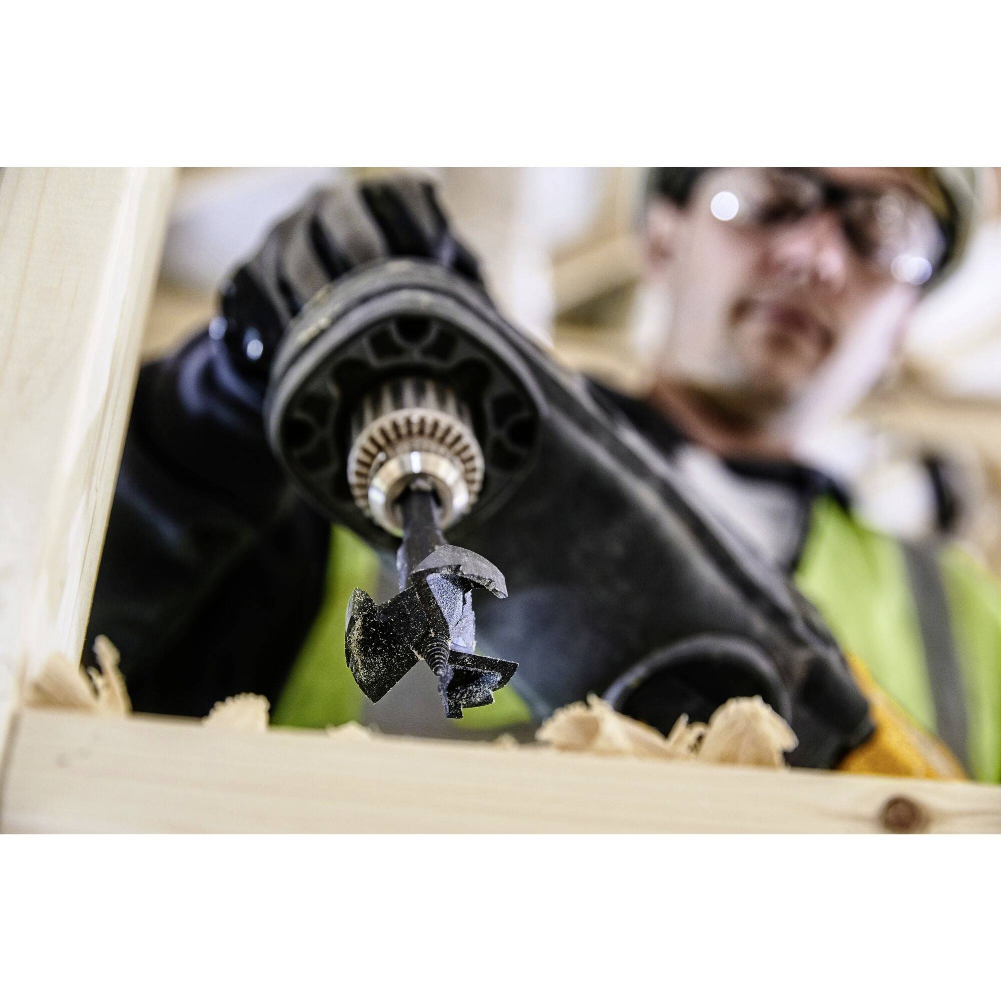 A construction worker in protective clothing is drilling a hole in a piece of wood with a cordless drill.