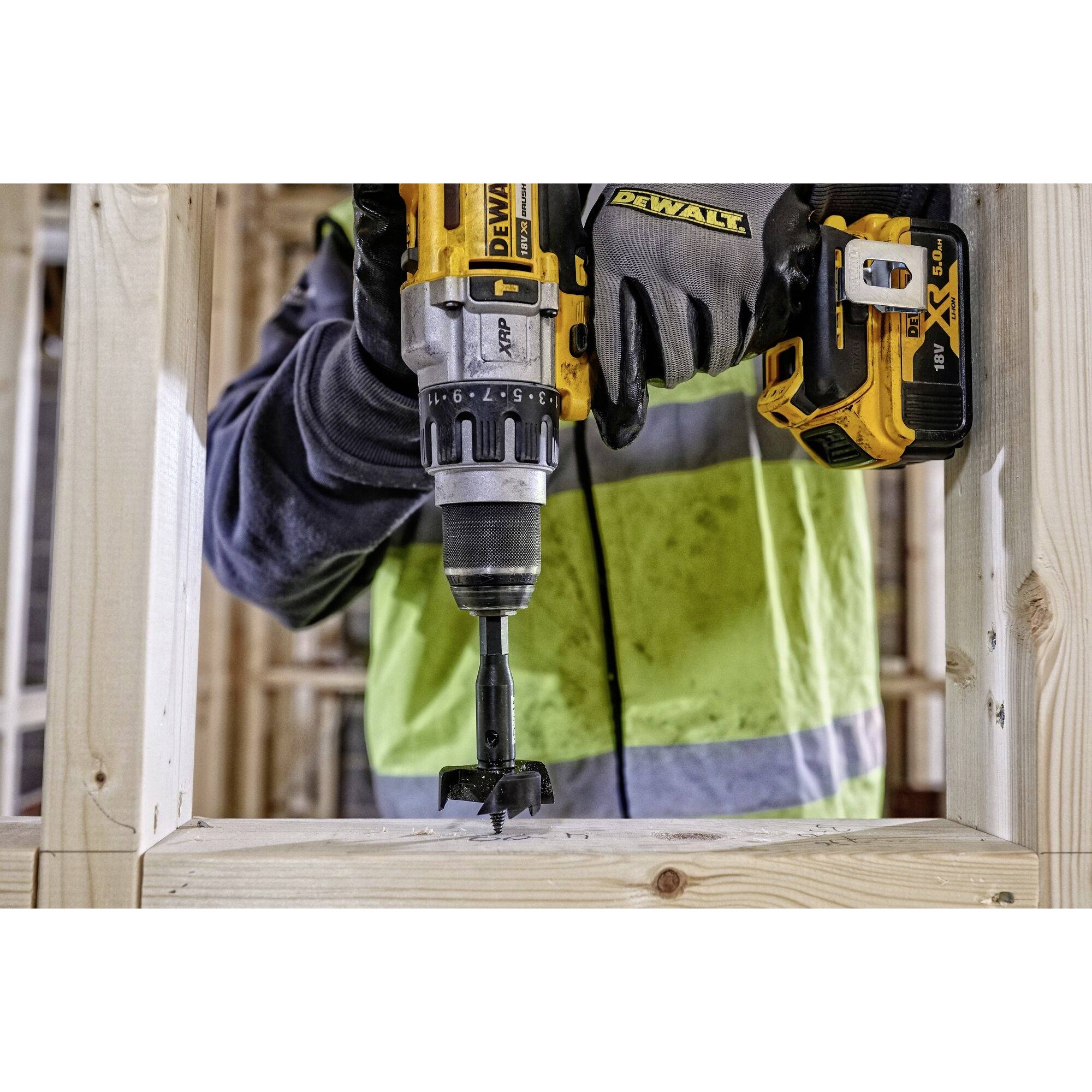 A person wearing a high-visibility safety vest is using a yellow, cordless drill to bore a hole in a wooden frame.