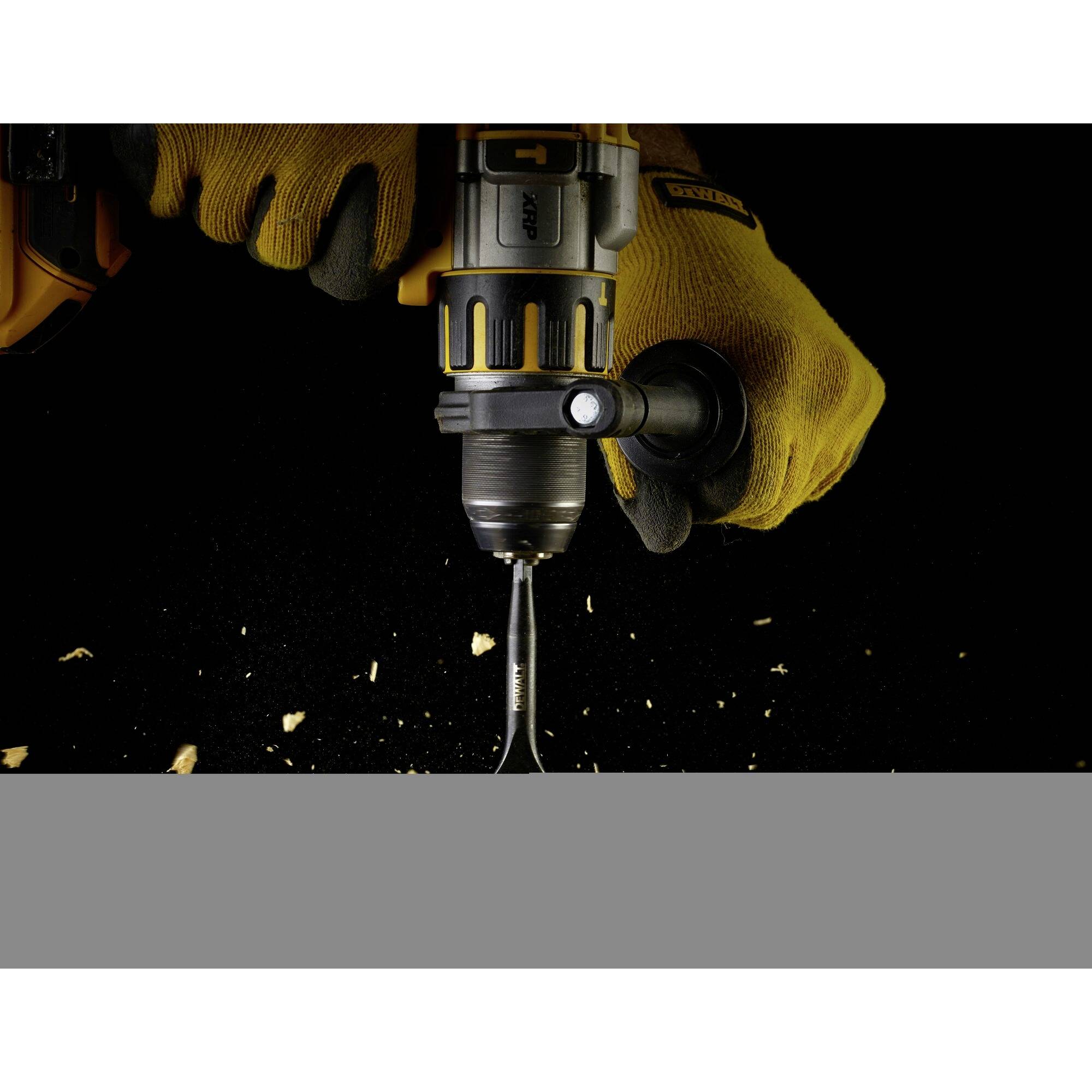 A person is wearing work gloves and holding a power drill. The drill bit is penetrating a piece of wood, with wood shavings flying around.