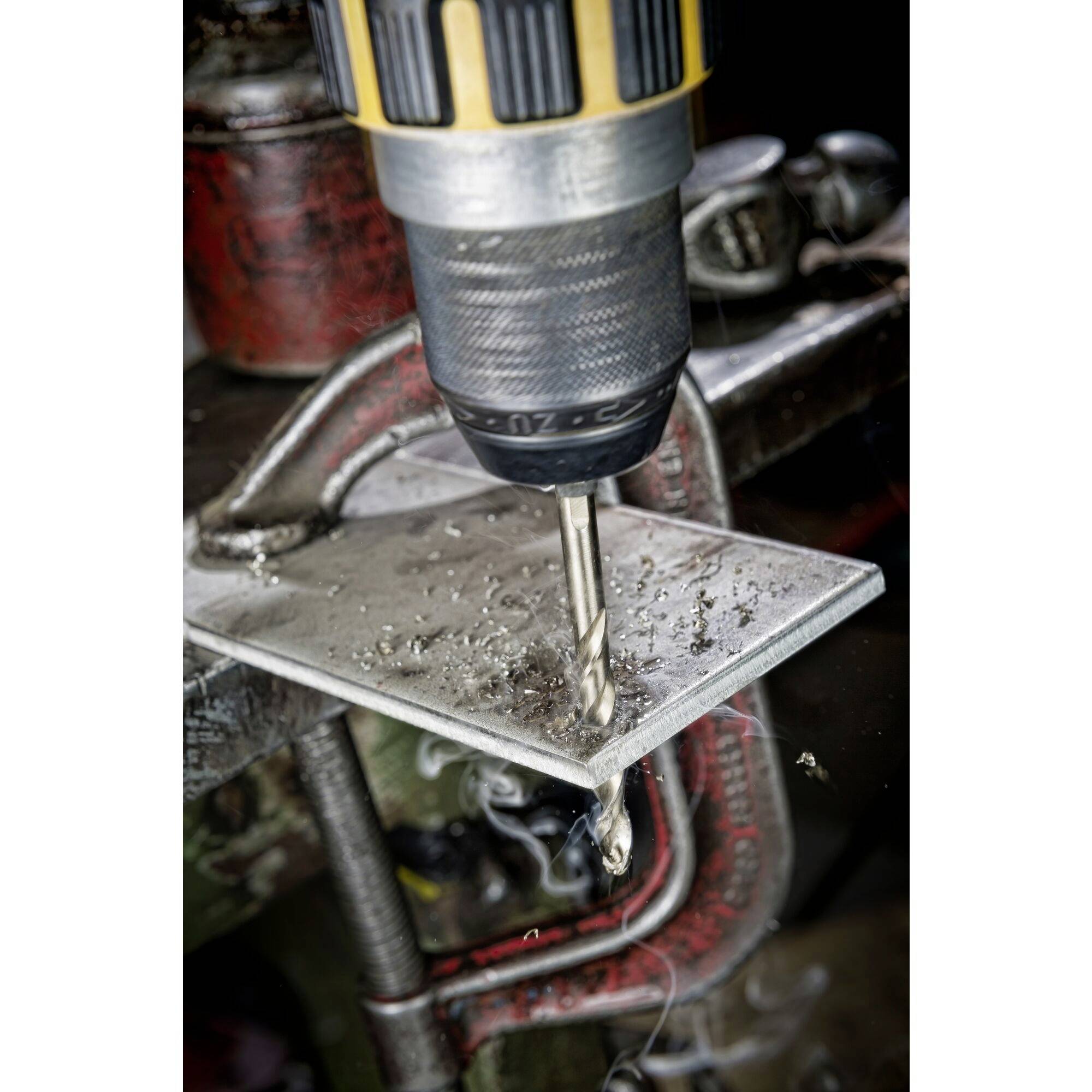 A drill with a metal drill bit is forming a hole in a metal piece secured in a vice. Metal shavings are visible.
