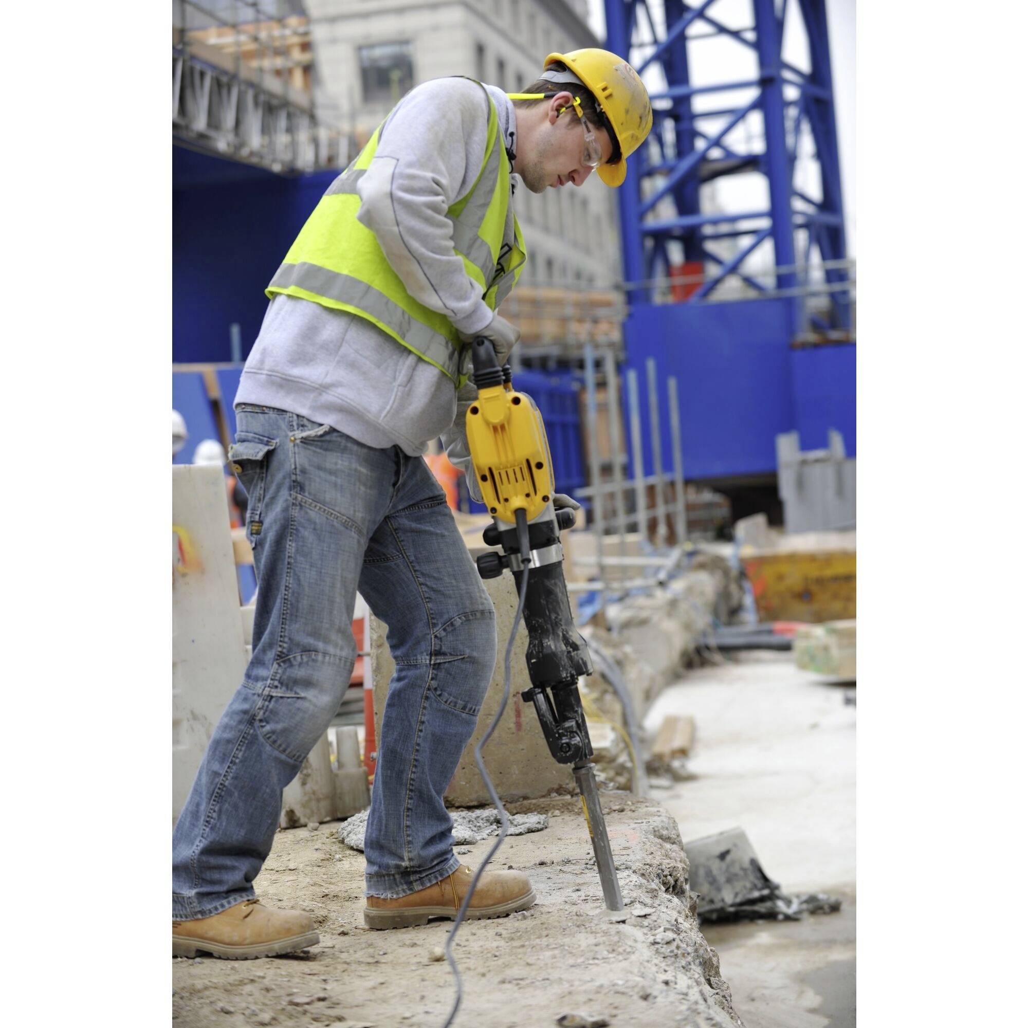 A person on a construction site is operating a pneumatic hammer, wearing a hard hat and a high-visibility vest. Scaffolding is visible in the background.