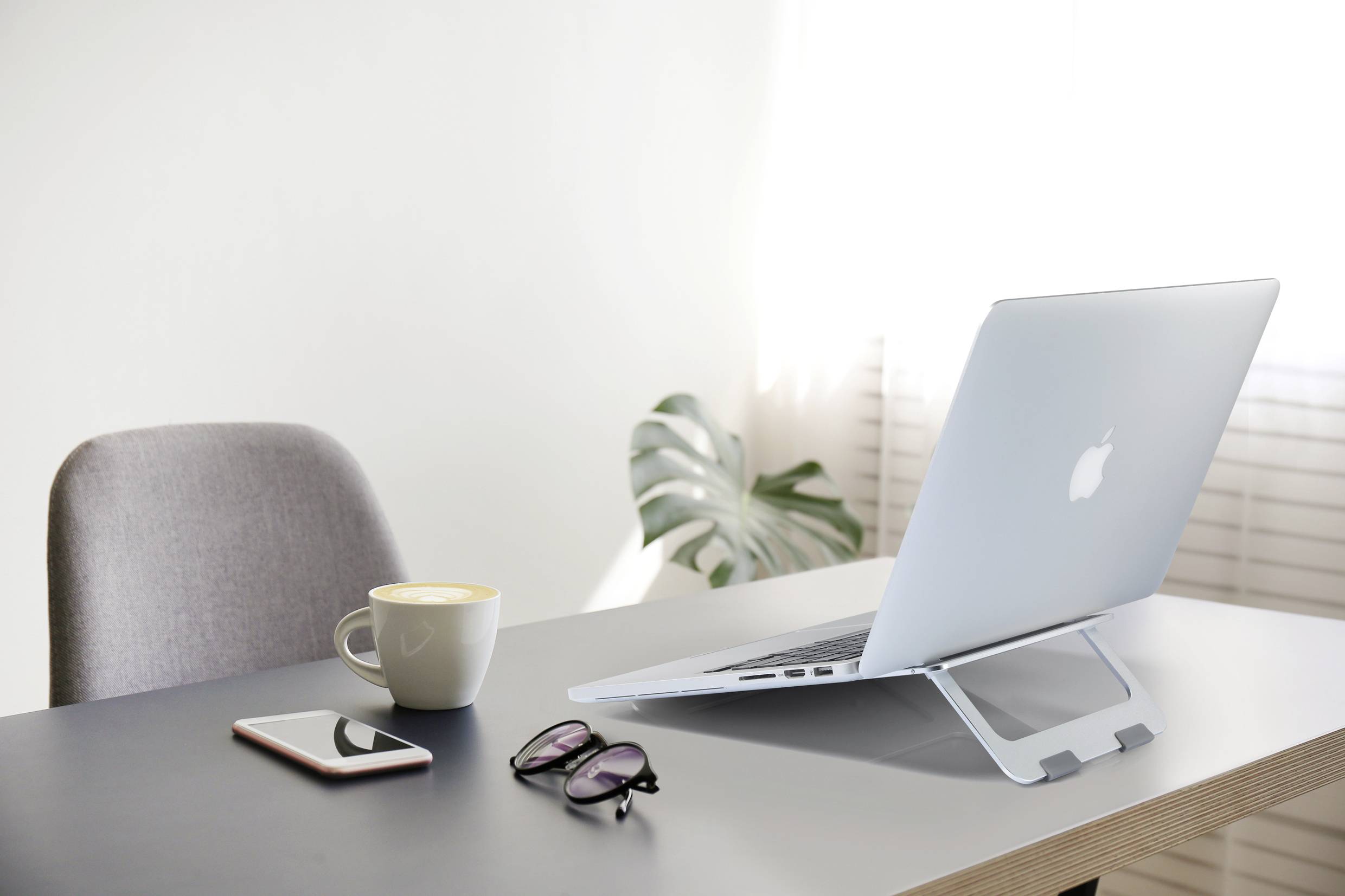 An open laptop on a stand on a desk, with a cup of coffee, a smartphone, and a pair of glasses beside it.