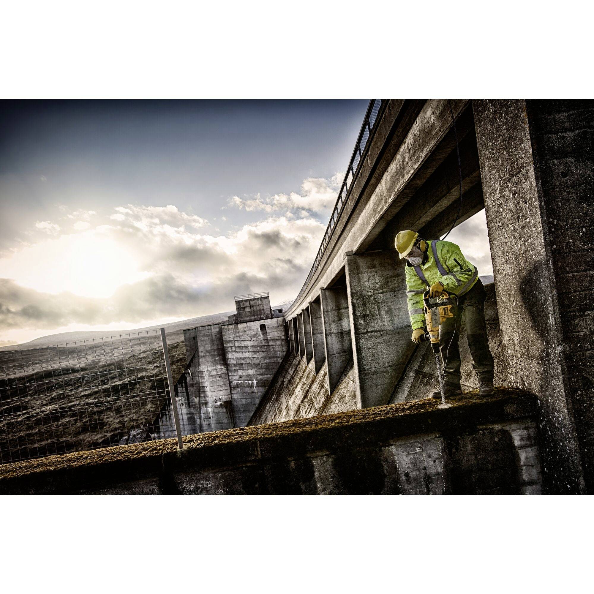 A person wearing protective equipment is working with an electrical tool on a dam at sunset. The environment appears industrial.