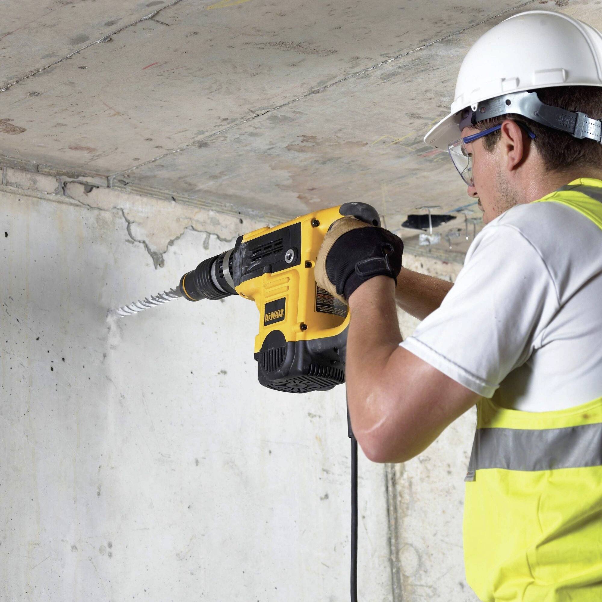 A person wearing a safety helmet and protective goggles is drilling into a concrete wall with a yellow drill.