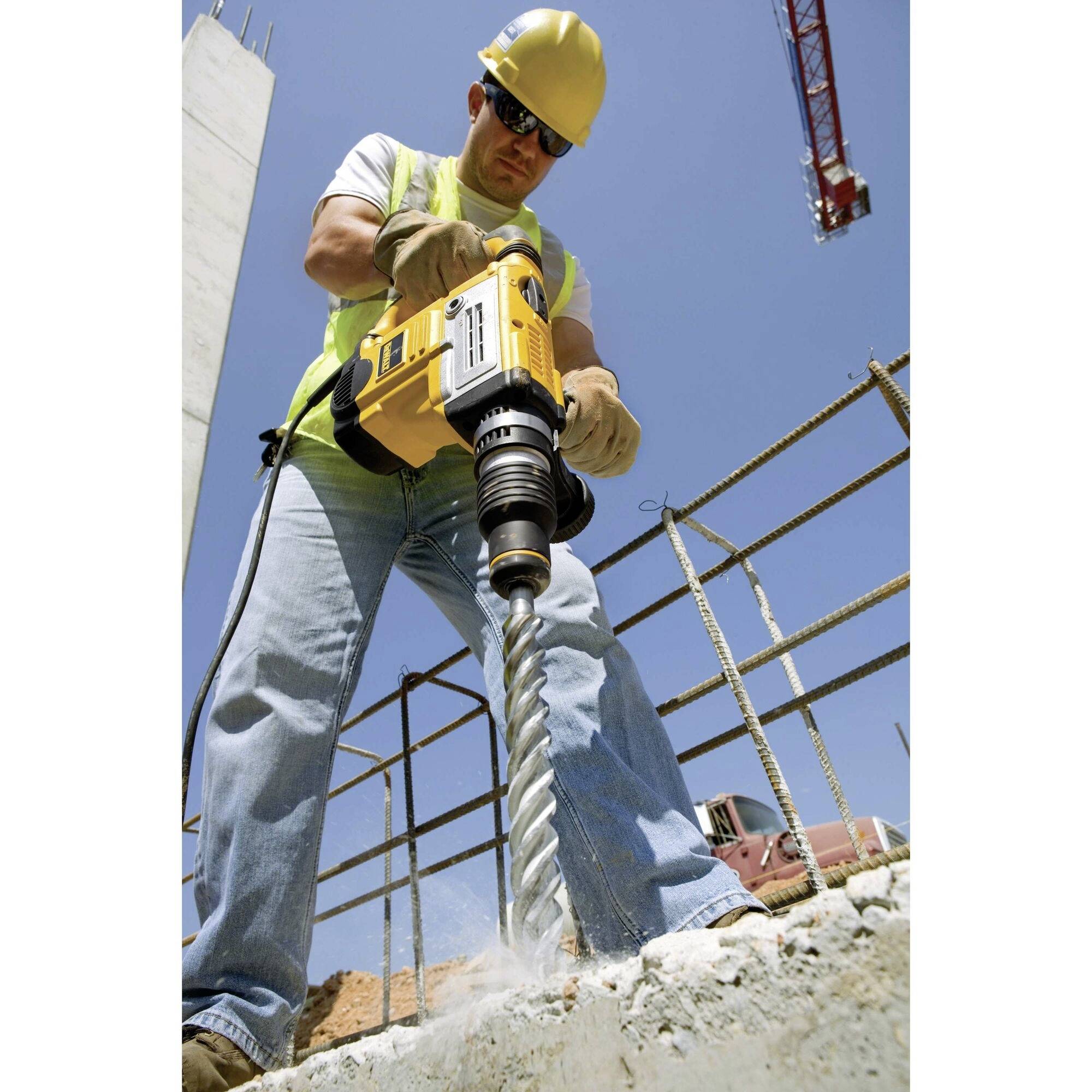 A construction worker wearing a hard hat and safety glasses is drilling into concrete with a yellow power drill, with a clear sky and a crane in the background.