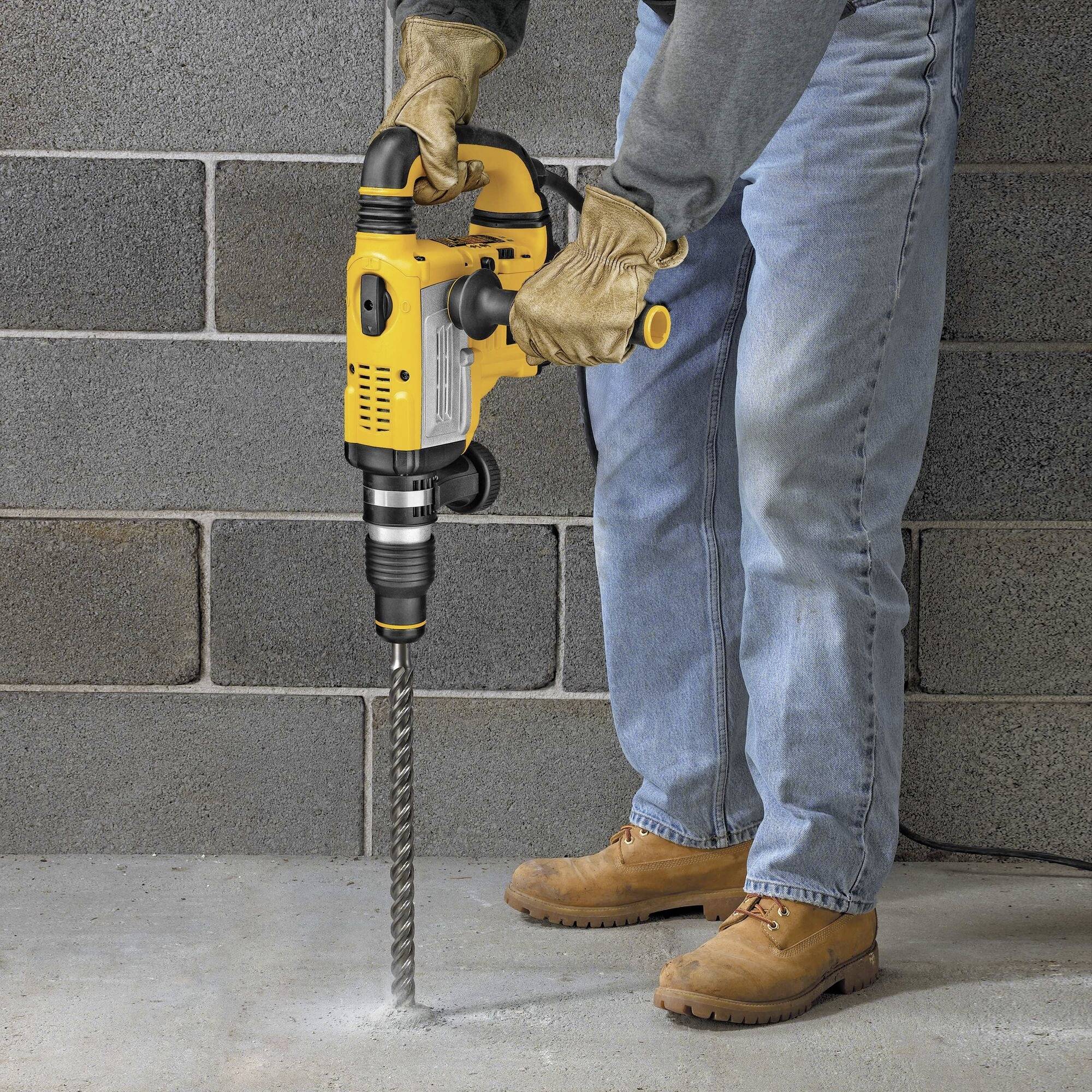 A person is drilling into a concrete wall with a yellow drill, wearing jeans, brown shoes, and gloves.
