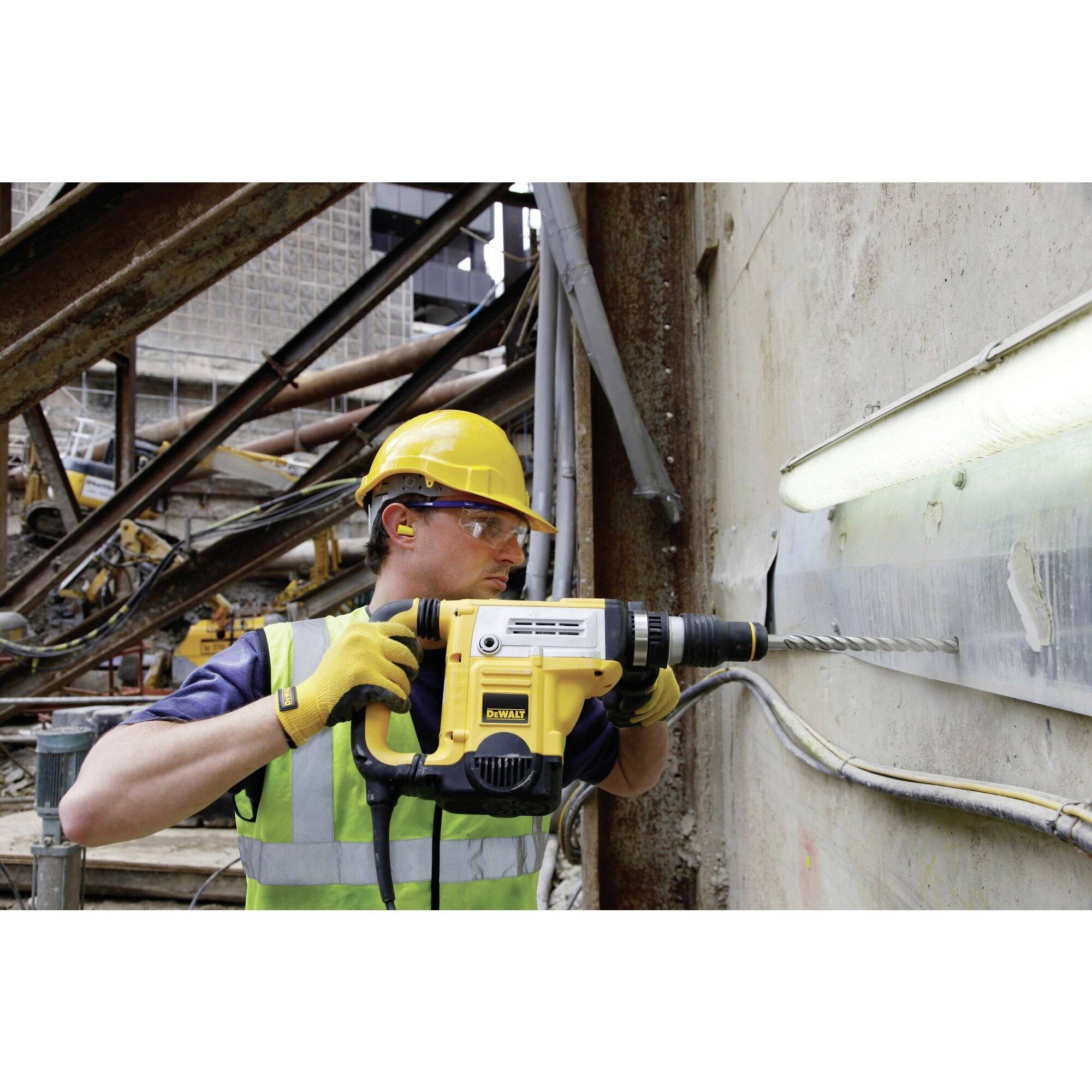 A construction worker in protective gear is drilling into a concrete wall on a building site using a yellow drill.