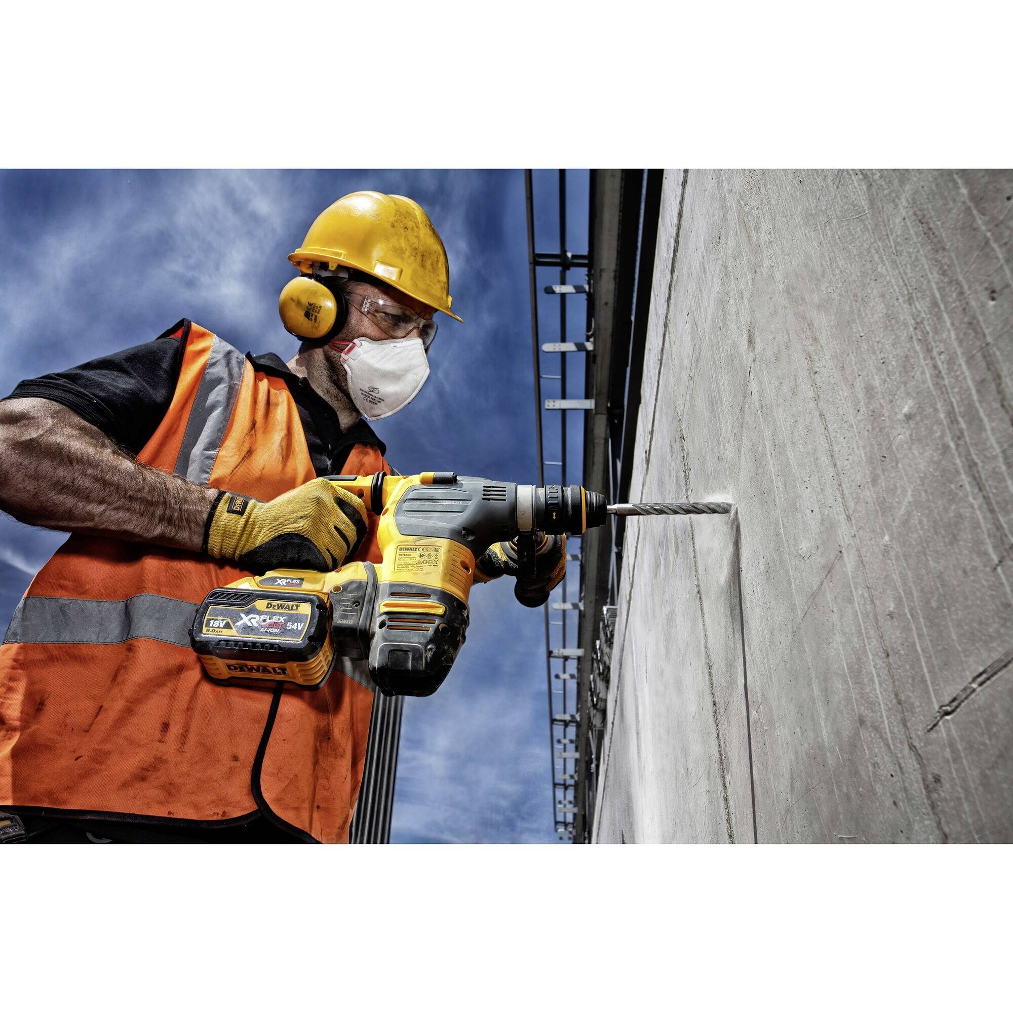 A construction worker in an orange high-visibility vest and hard hat is drilling a hole in a concrete wall using an electric drill.
