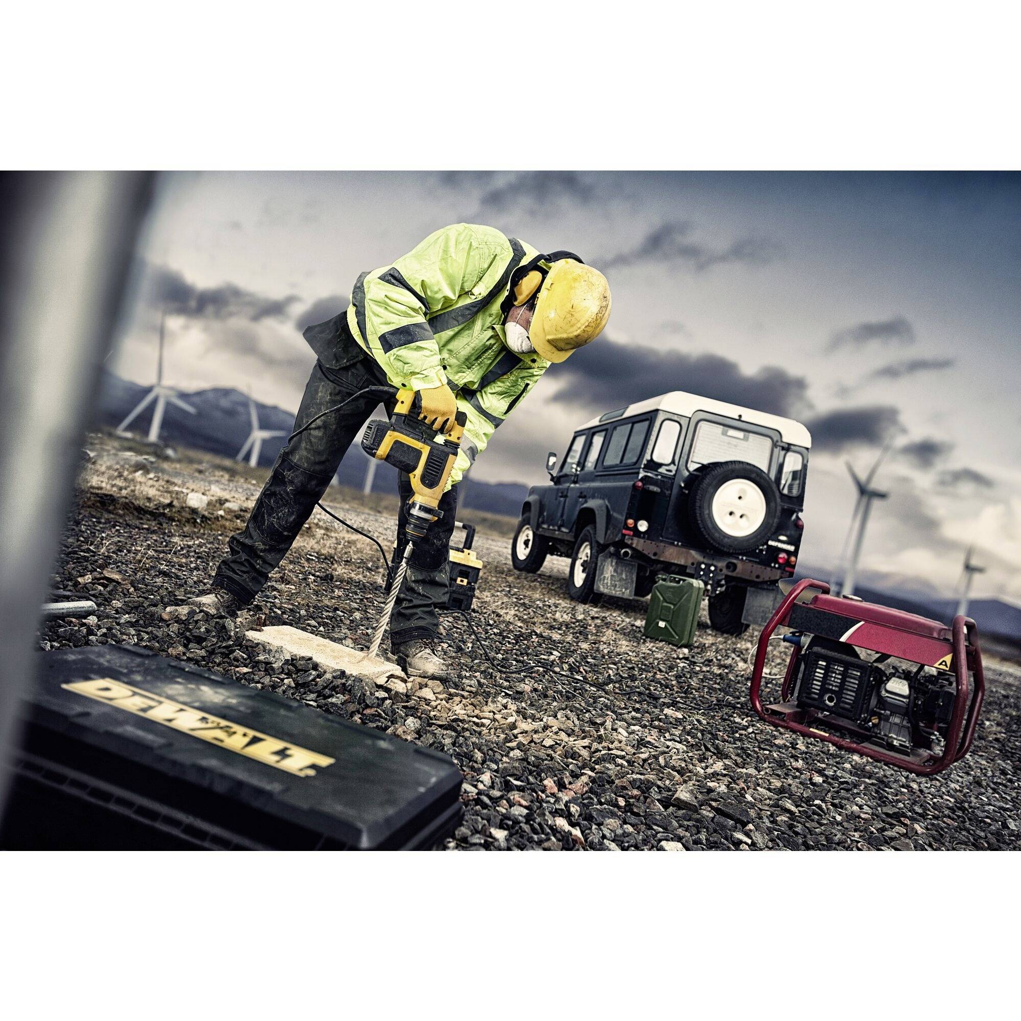 A worker in protective clothing is drilling into the ground outdoors. Wind turbines, an off-road vehicle, and a generator are visible in the background.