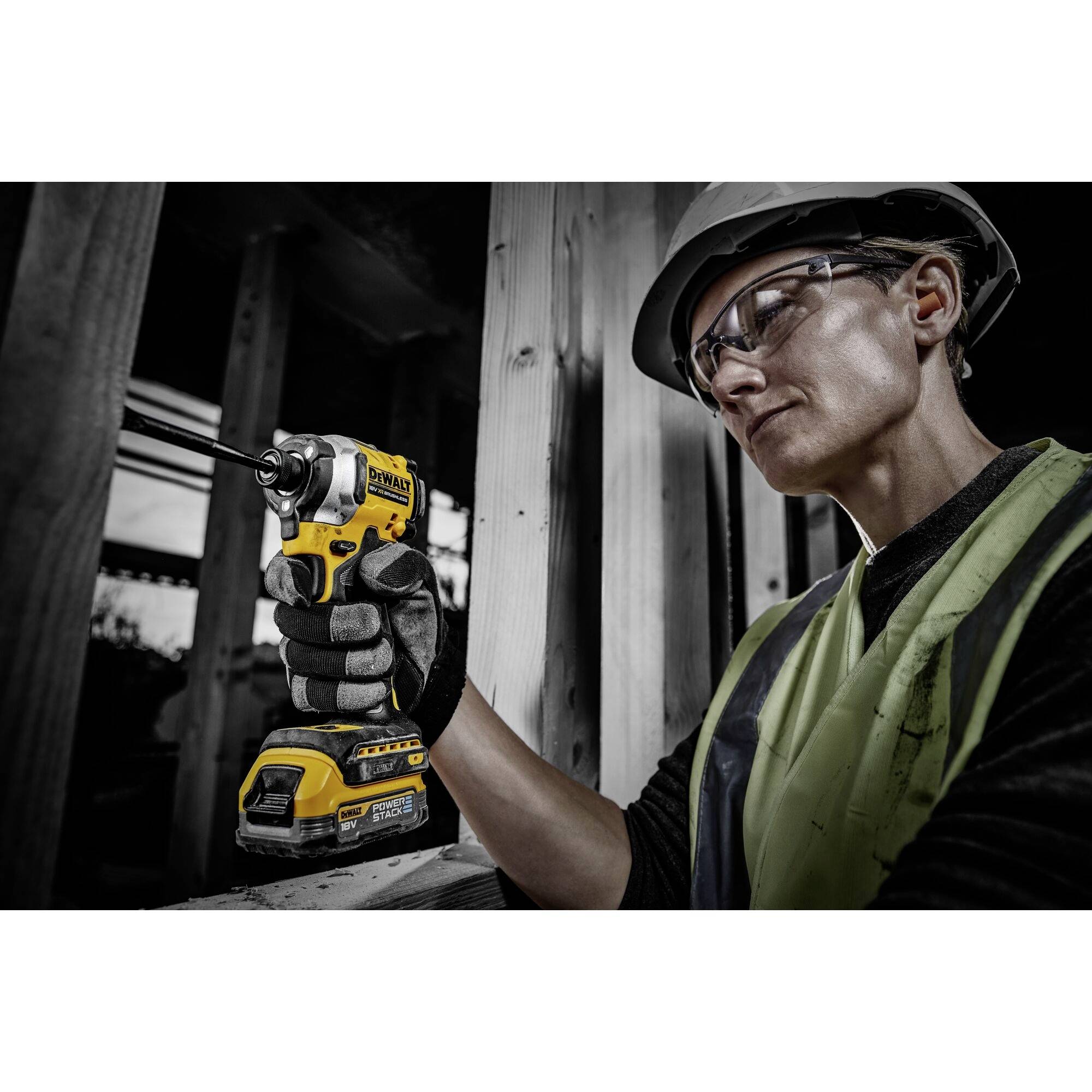 A worker wearing safety glasses and a hard hat is holding a yellow cordless drill and working on wood in a construction site environment.
