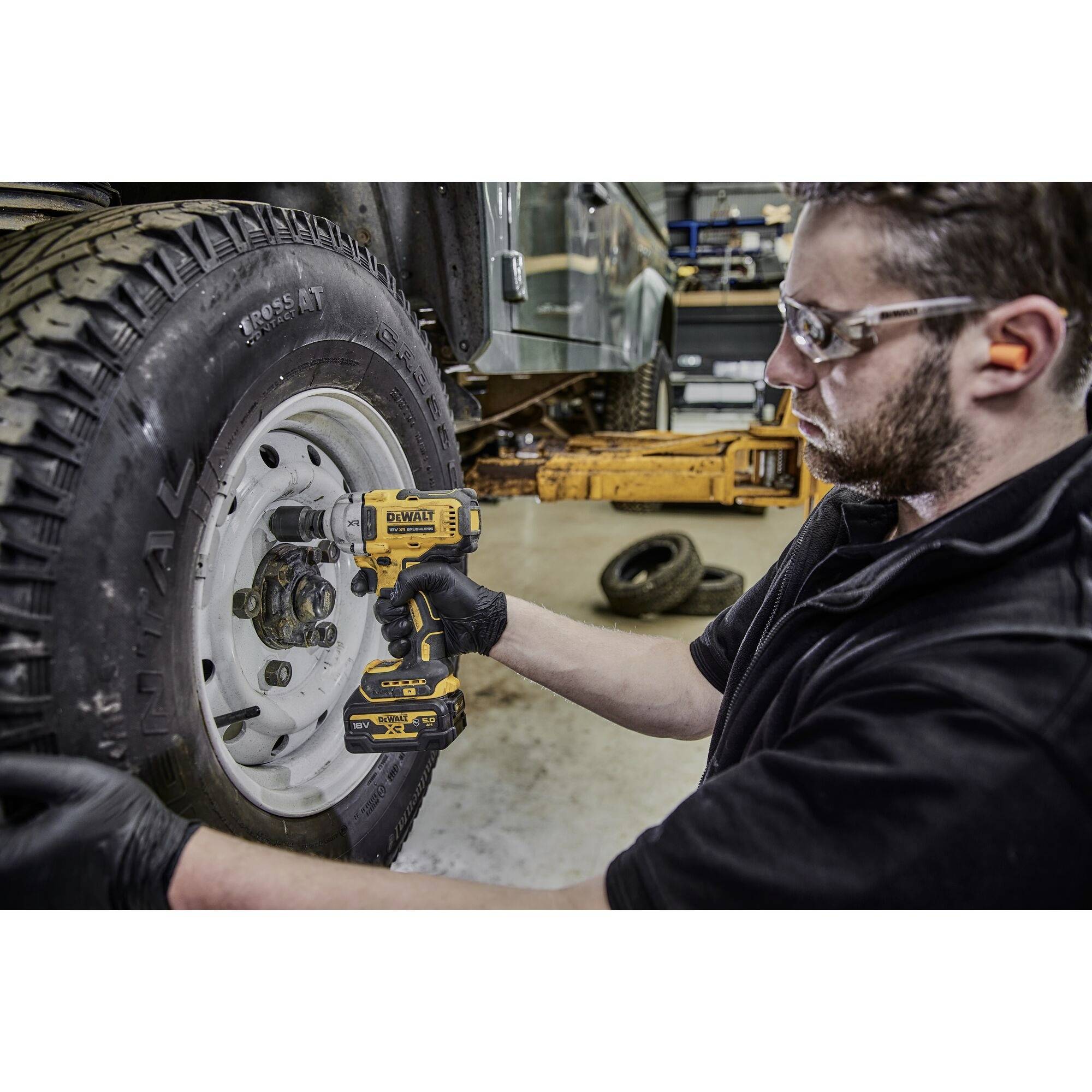 A man is using an electric torque wrench to loosen the wheel nuts of a vehicle. He is wearing safety glasses and gloves.