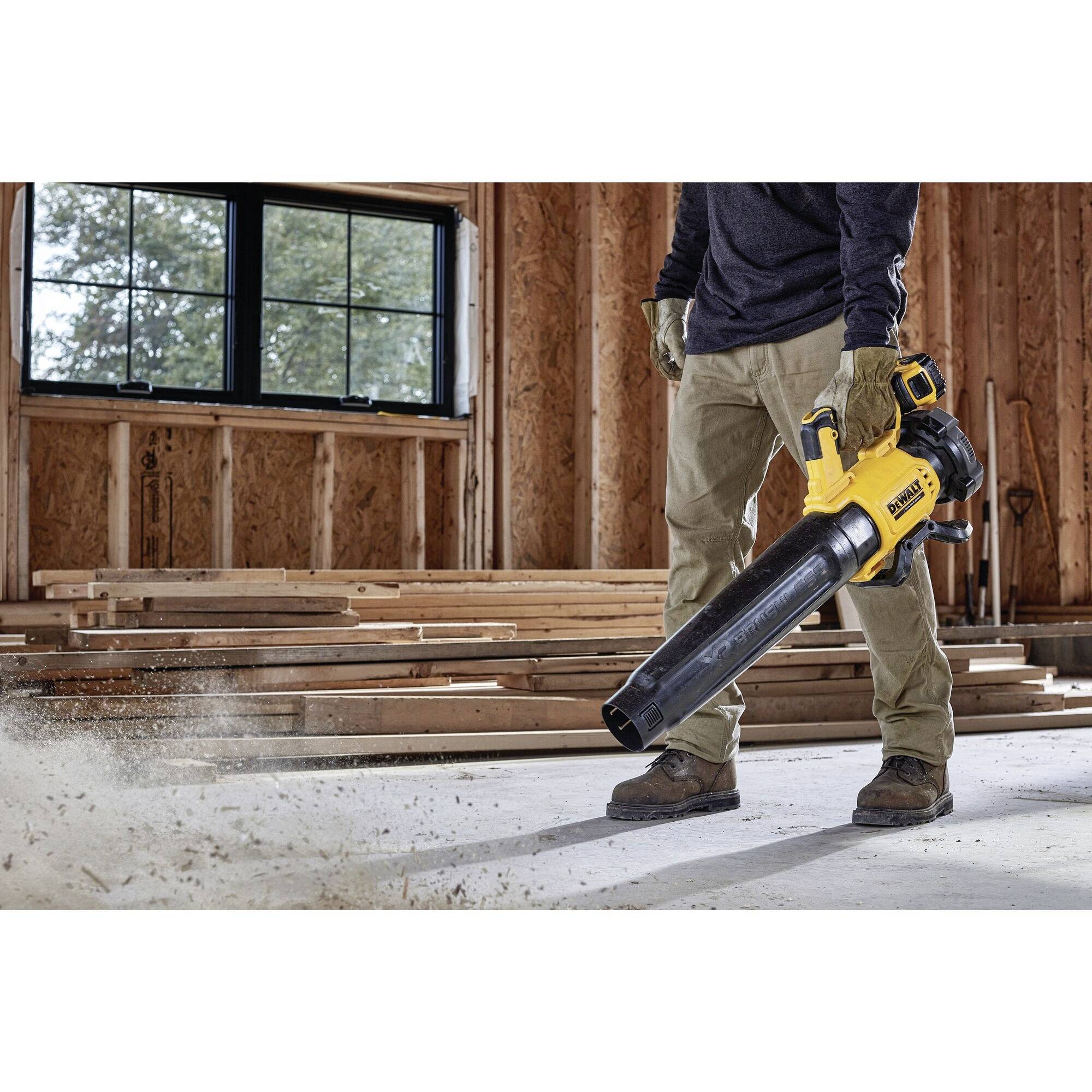 A person is using a yellow leaf blower in a workshop with wood stacks in the background. The floor is dusty.