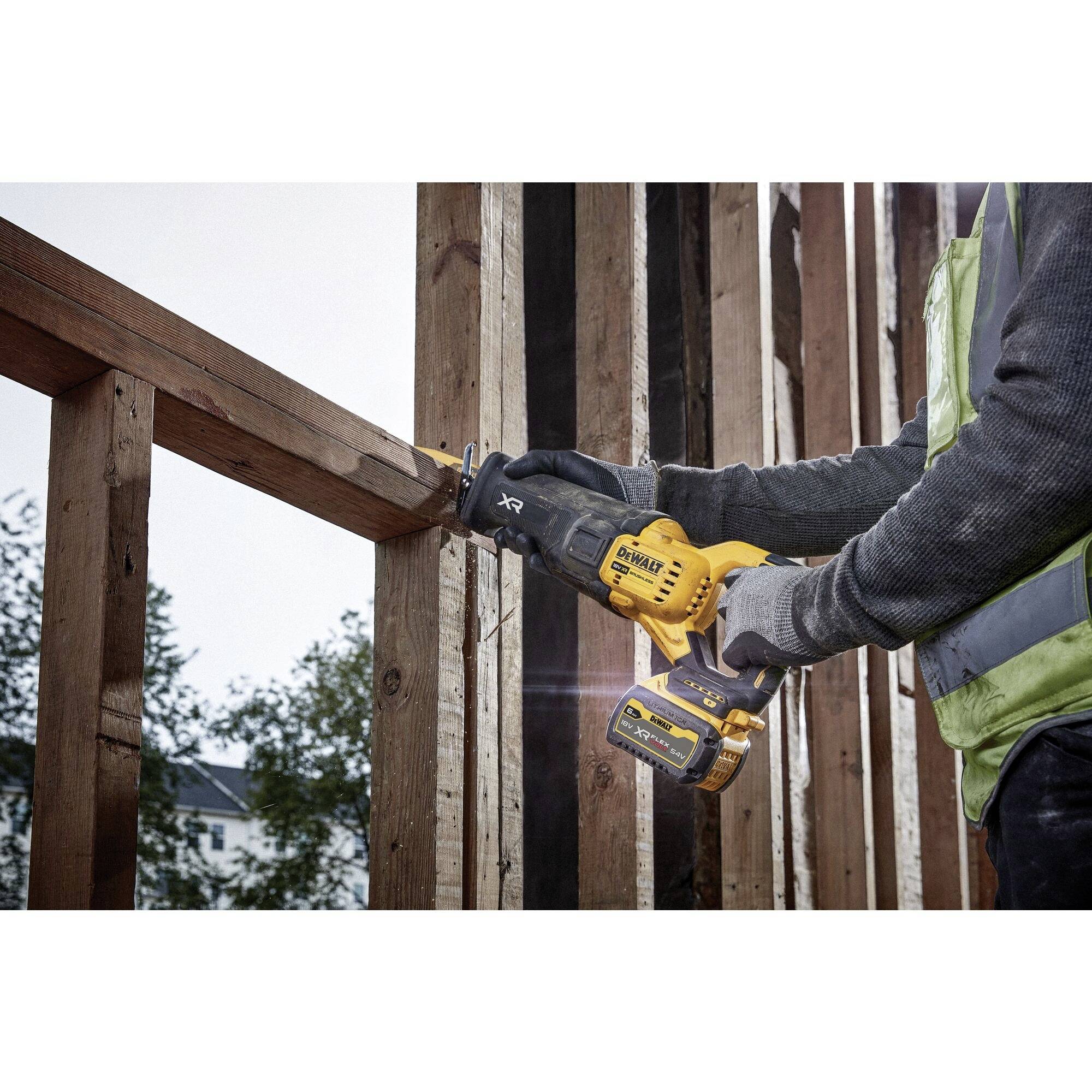 'Worker sawing wooden frame with cordless reciprocating saw, wearing safety vest. Background shows incomplete building construction.'