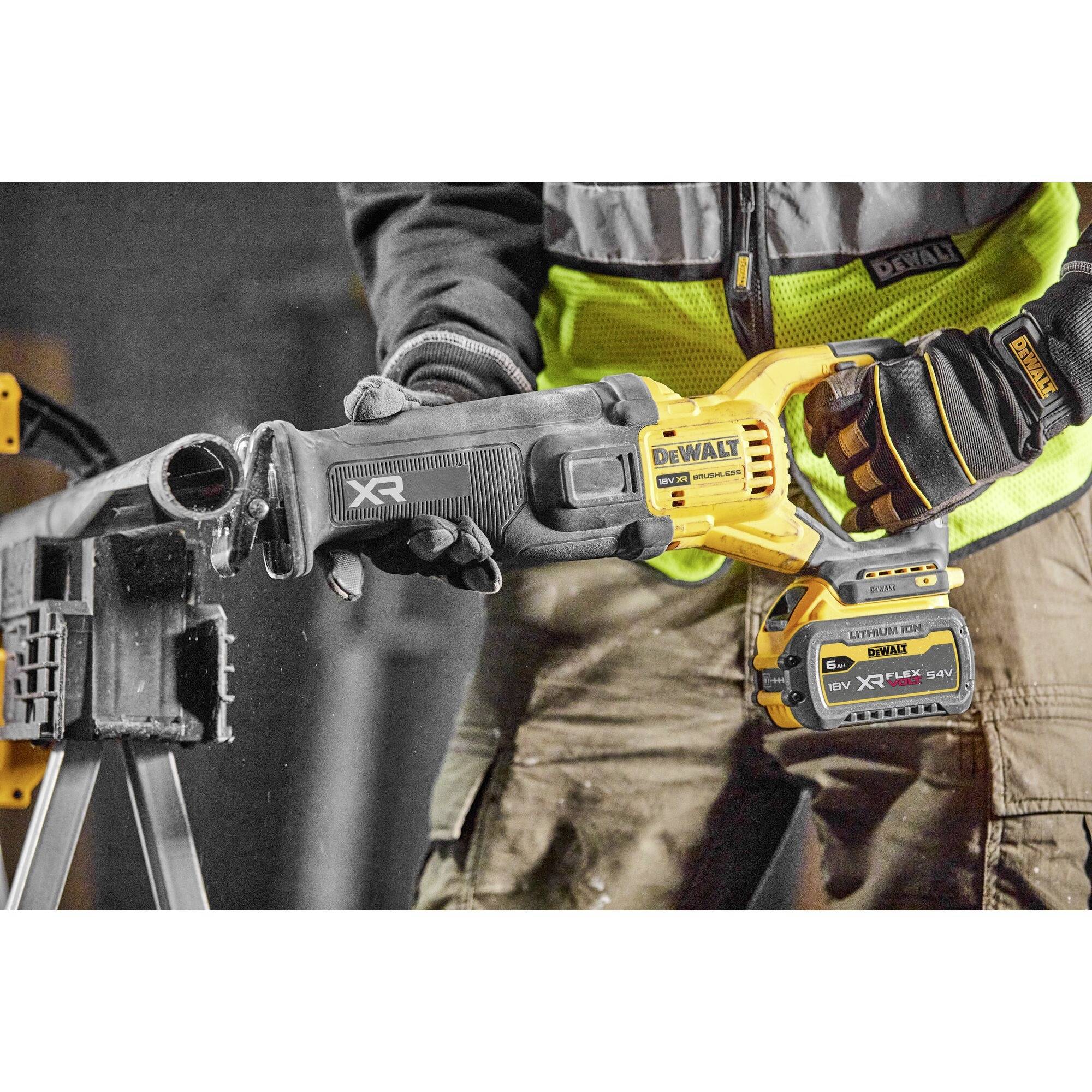 A person is using a yellow cordless reciprocating saw to cut through a pipe. They are wearing protective workwear with gloves.