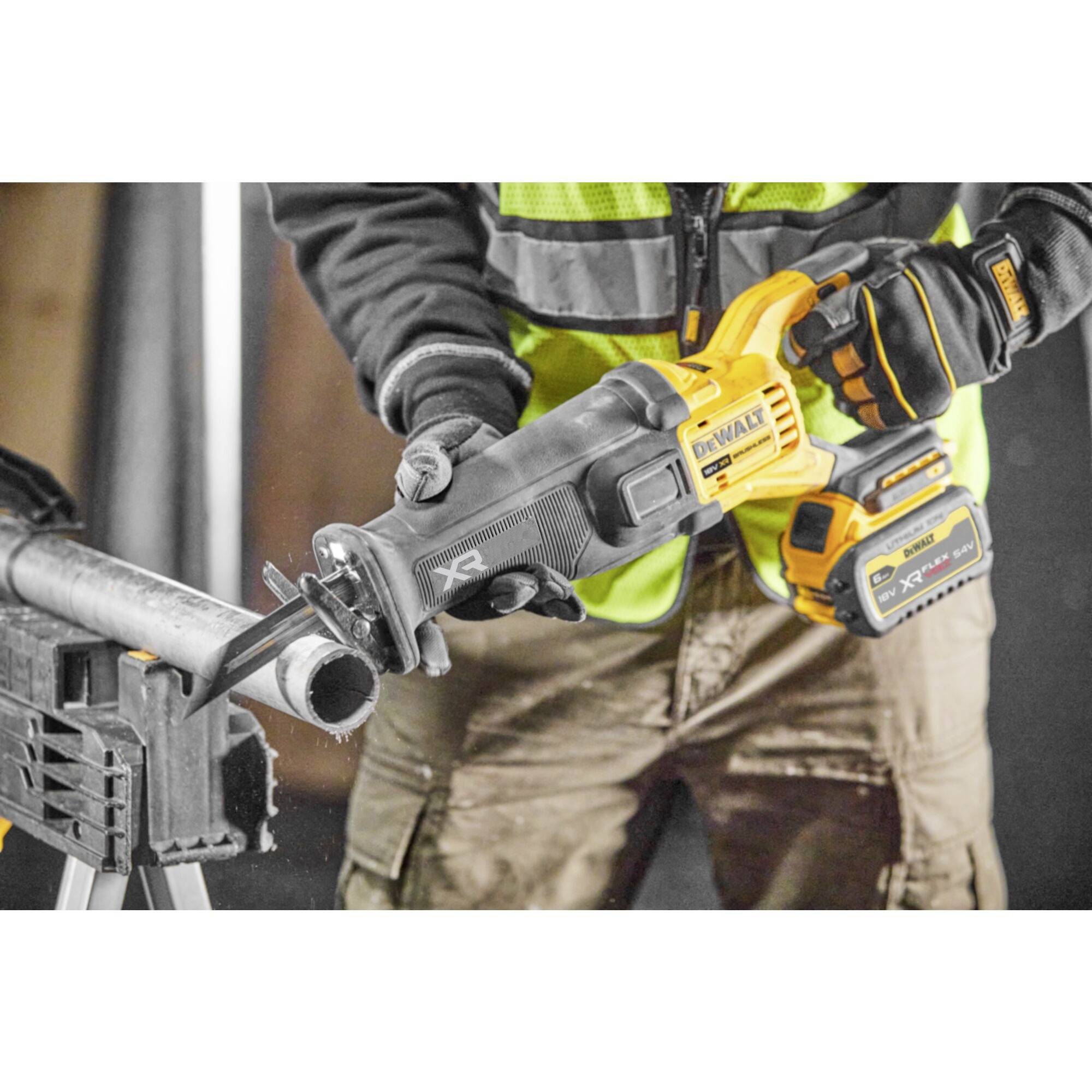 A person wearing a high-visibility safety vest is using a cordless reciprocating saw to cut a metal pipe on a workbench.