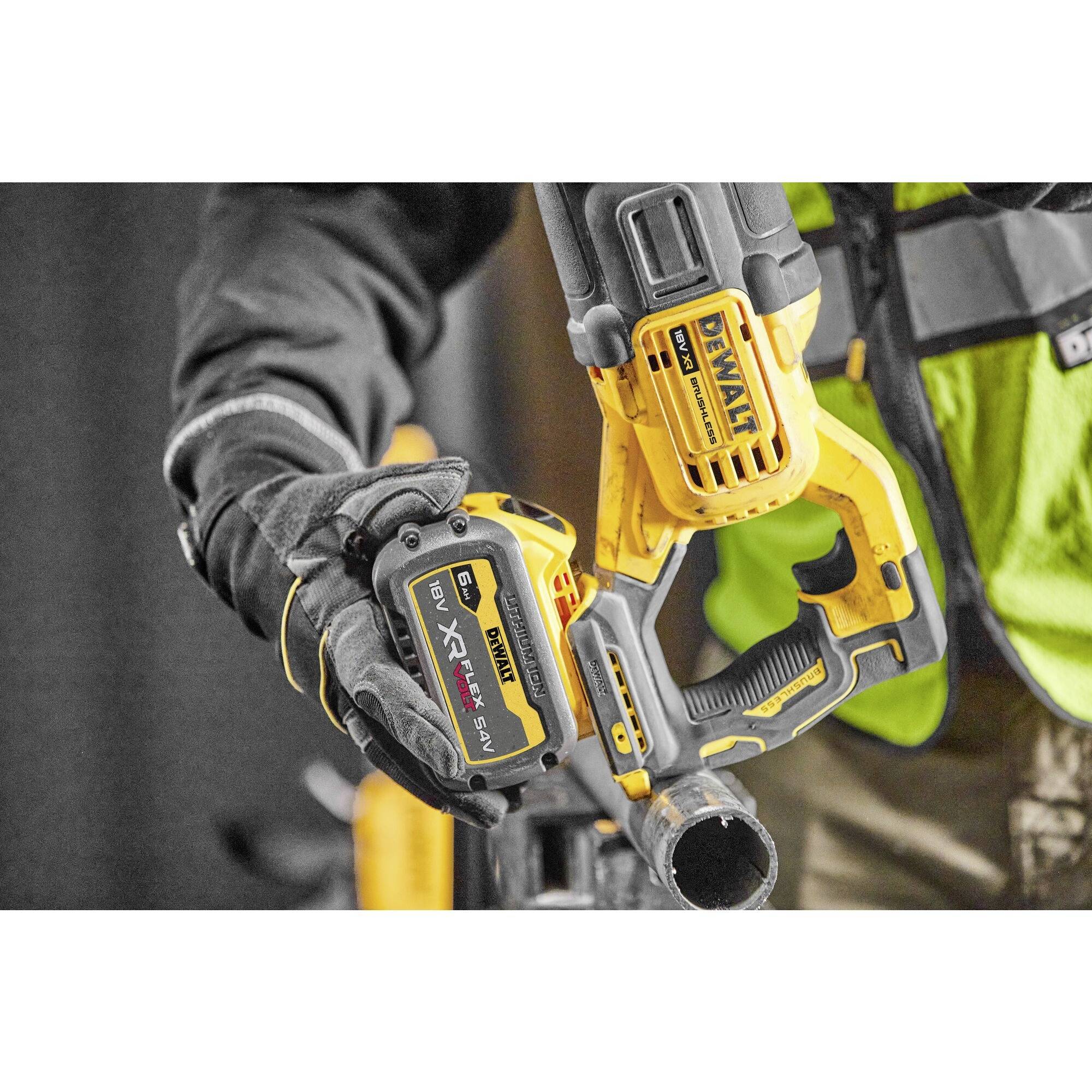A person is holding a yellow, cordless saw. The person is wearing work gloves and a high-visibility safety vest.