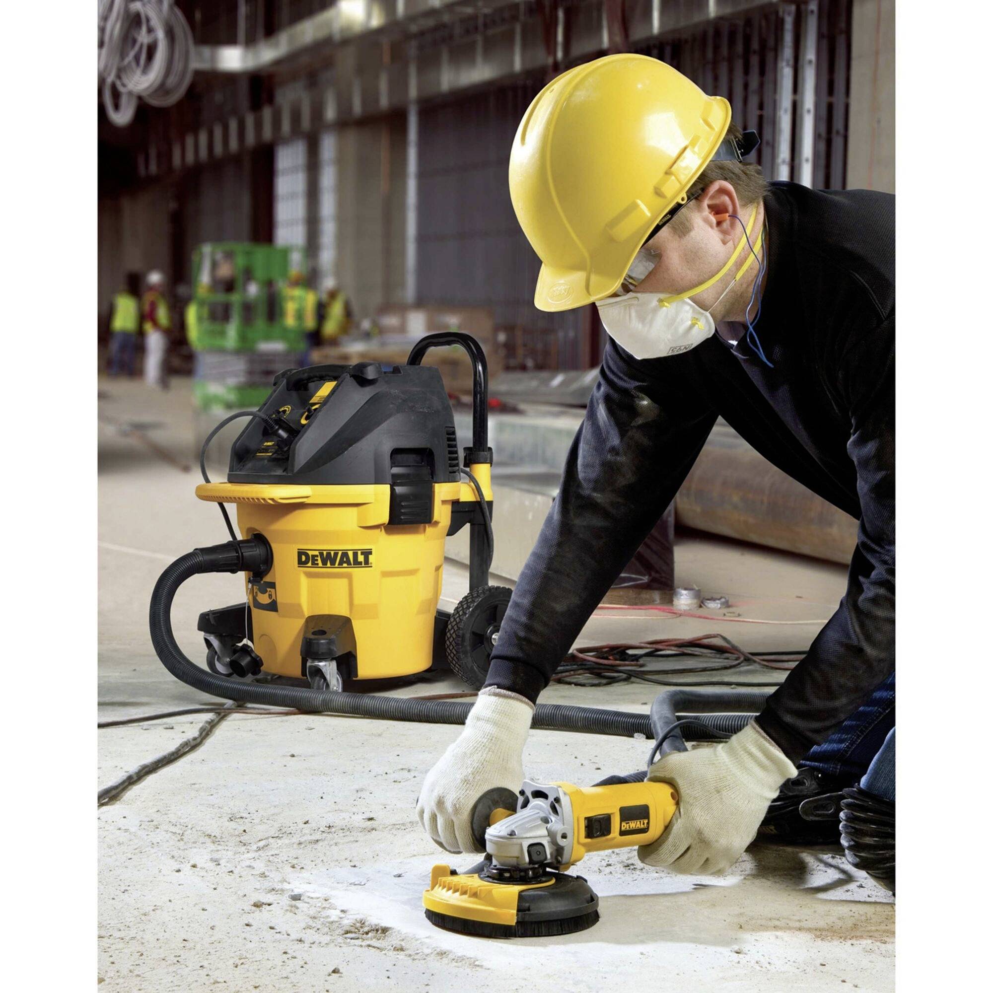 A worker is wearing protective clothing and using an electric tool to grind a concrete surface, with an industrial vacuum cleaner in the background.