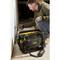 A man is working on a wall with a screwdriver, with a tool bag containing various tools standing nearby.