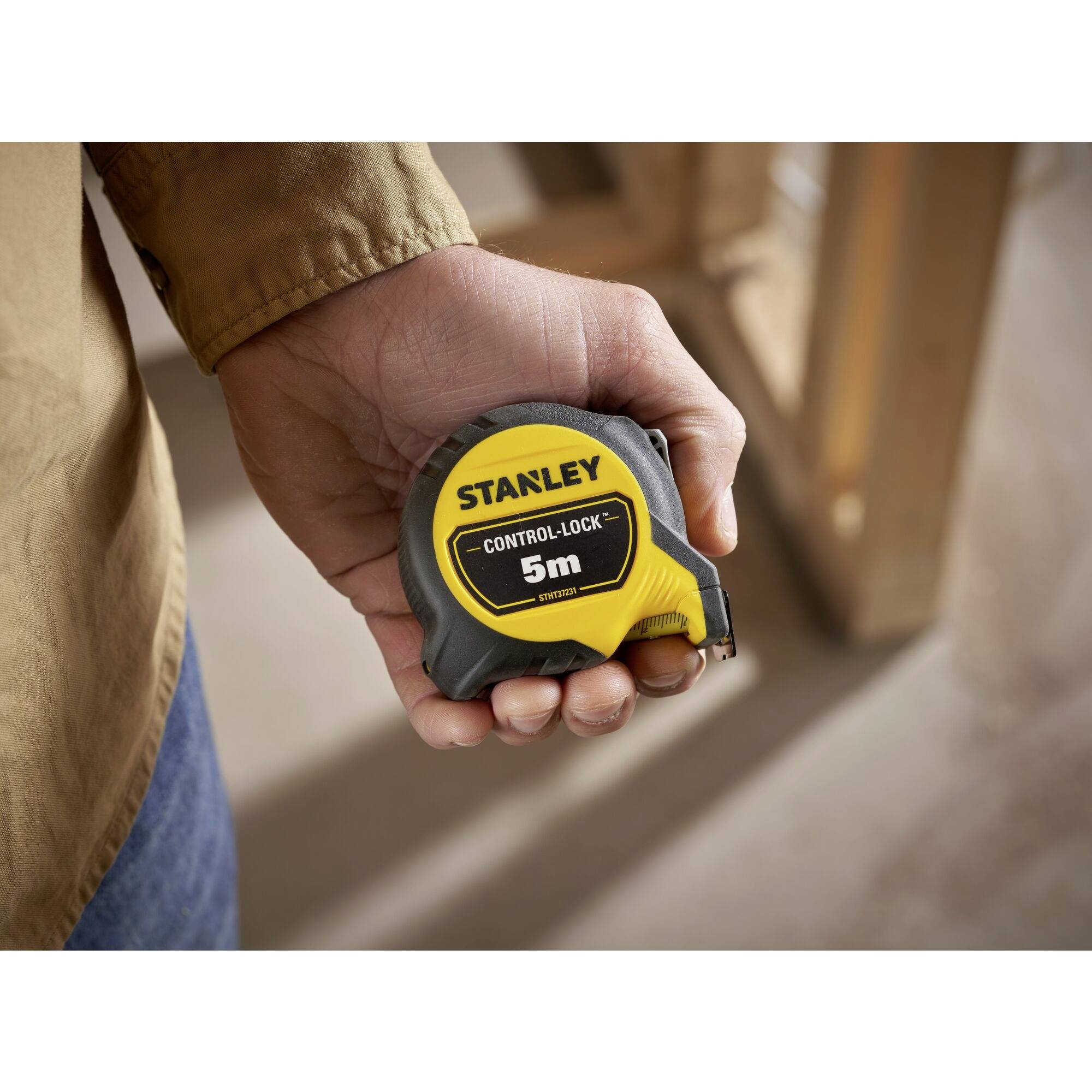 A hand is holding a 'Stanley' tape measure, 5 metres long. The surroundings suggest a construction site or workshop.