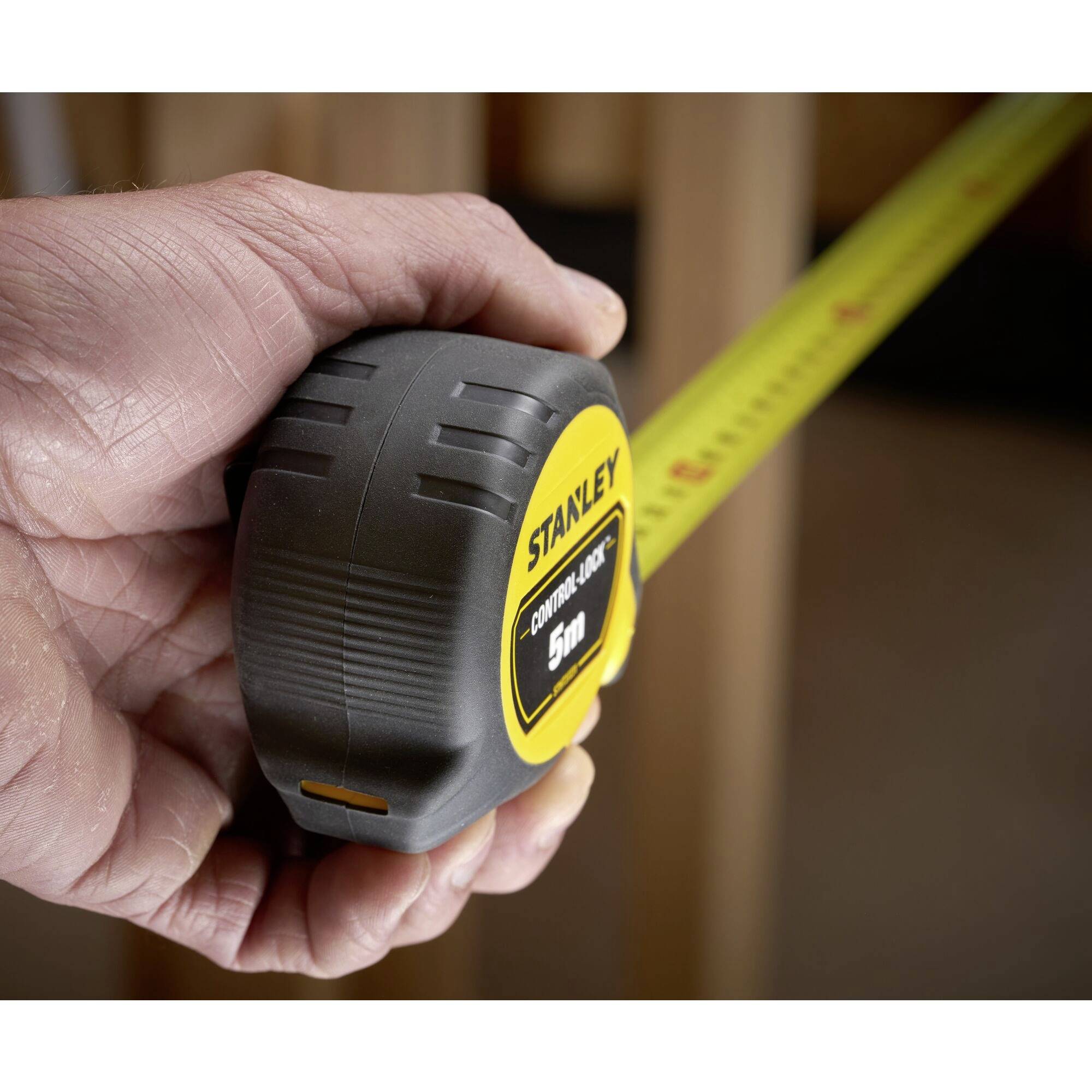 A hand is holding a yellow tape measure with the inscription 'STANLEY 5m', which is stretched out against a wooden post.