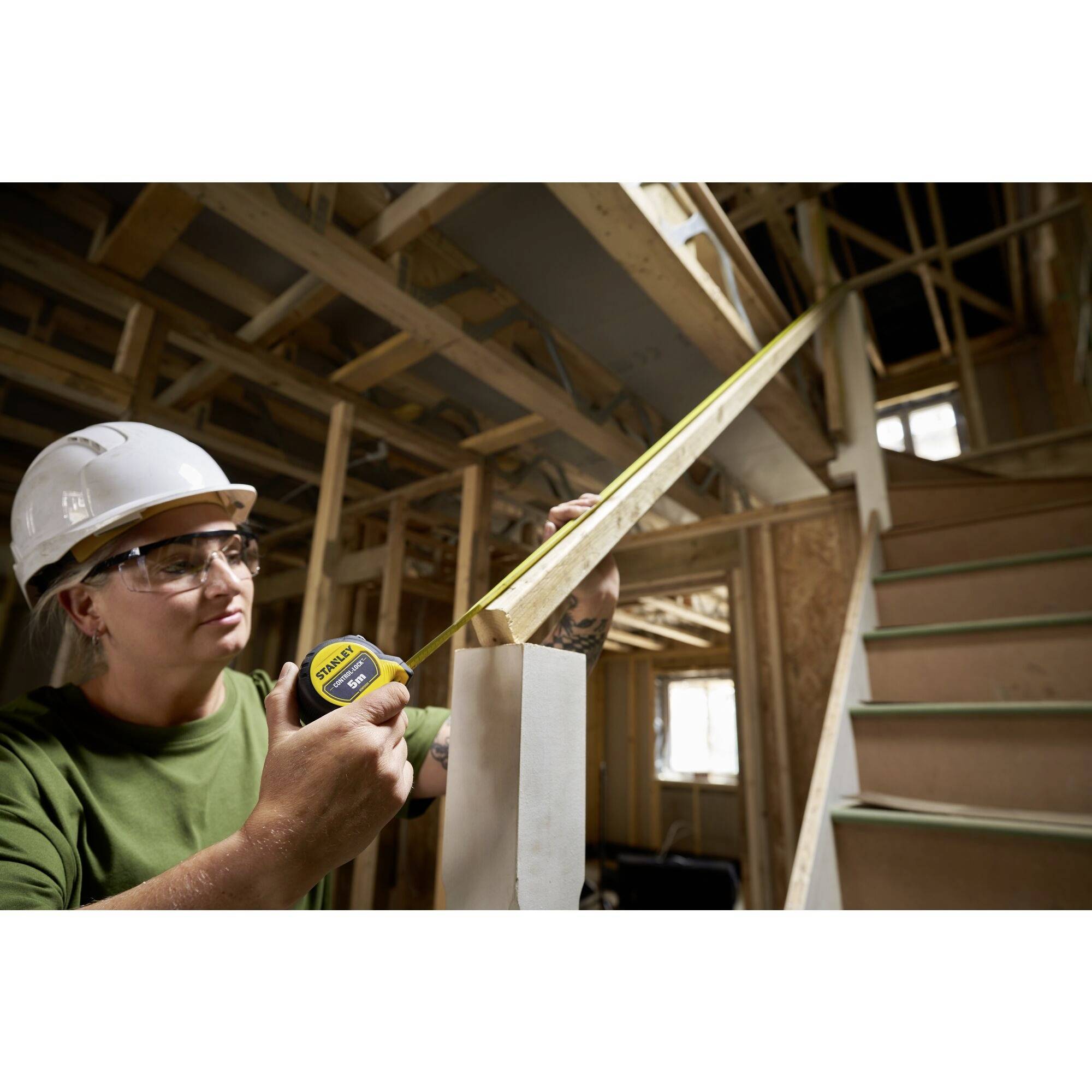 A person wearing a hard hat is measuring a wooden batten with a tape measure in a building under construction.