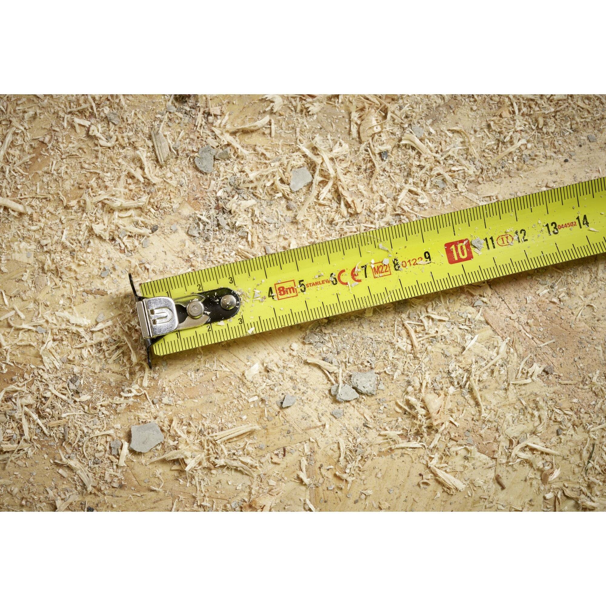 A yellow tape measure lies on a wooden surface with wood shavings, showing approximately 14 cm in length.