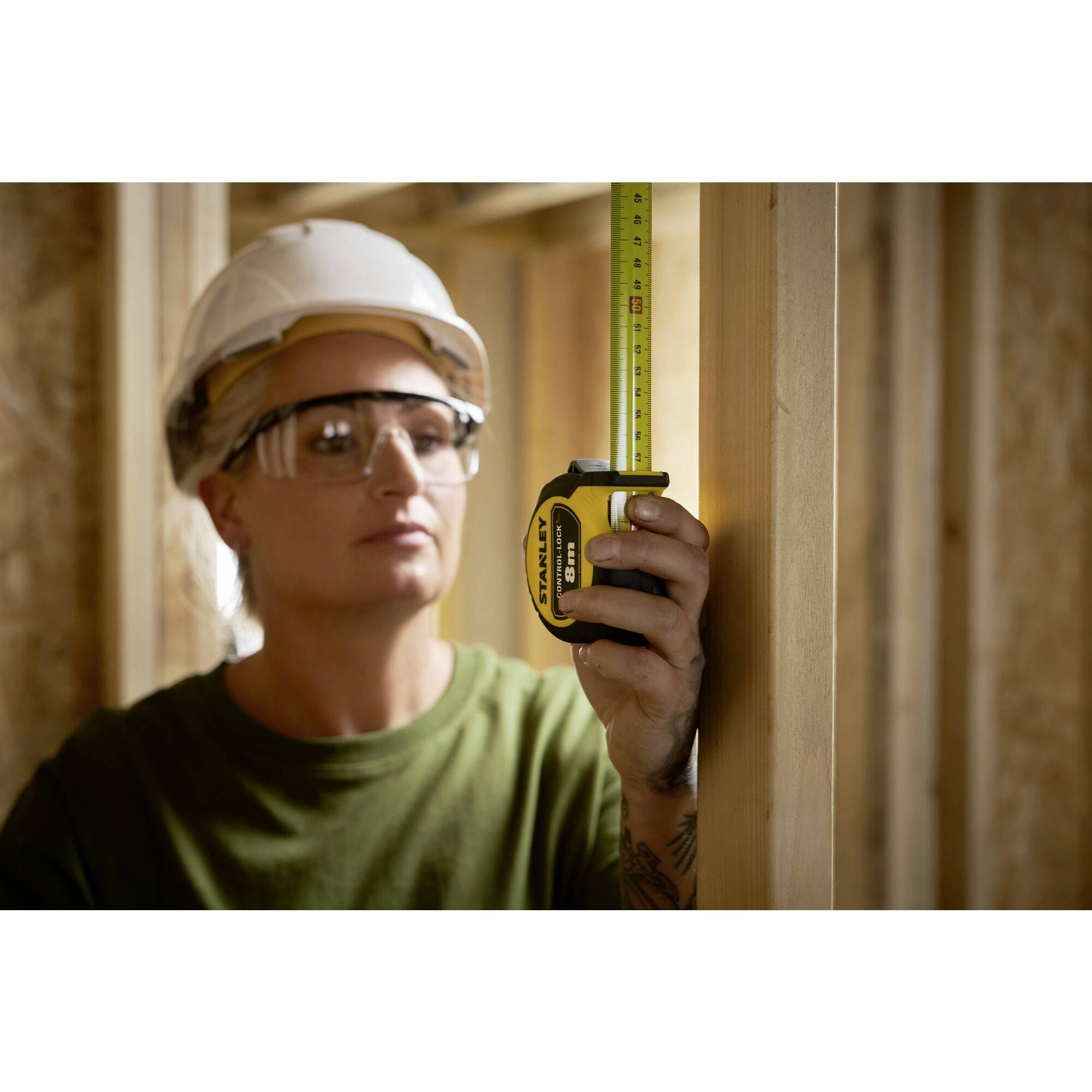 A person wearing a hard hat and safety glasses is measuring wooden frames with a tape measure in an unfinished building.