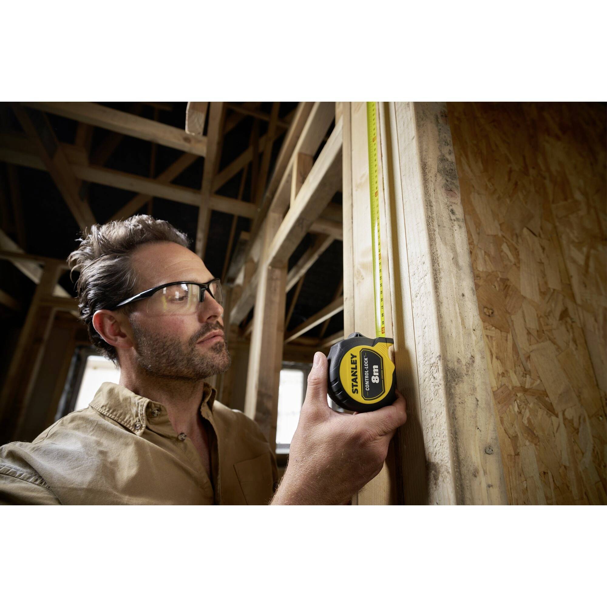 A man is measuring a wooden structure in a building using a tape measure. He is wearing safety glasses and working carefully.