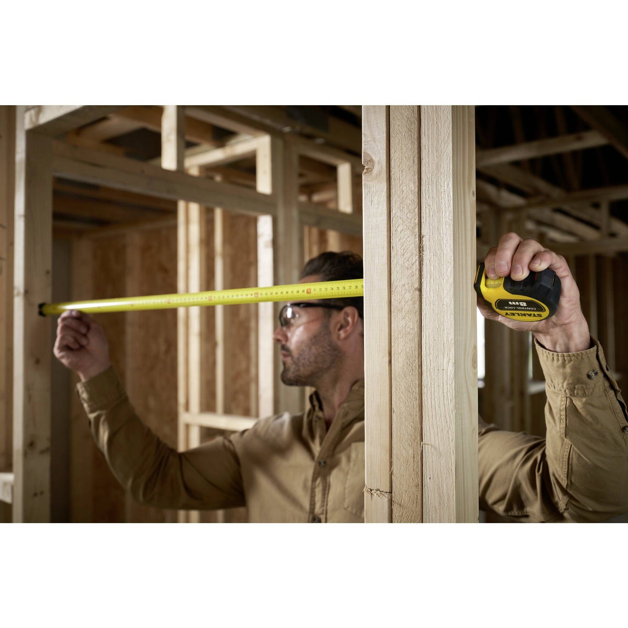 A man is measuring the width of a wooden frame with a tape measure in an unfinished room. The surroundings show ongoing construction work.