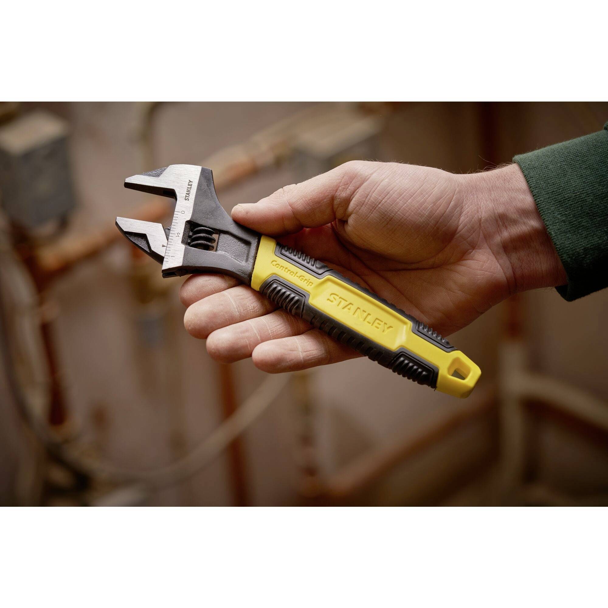 A hand is holding an adjustable spanner in a workshop environment. The handle is yellow and black.