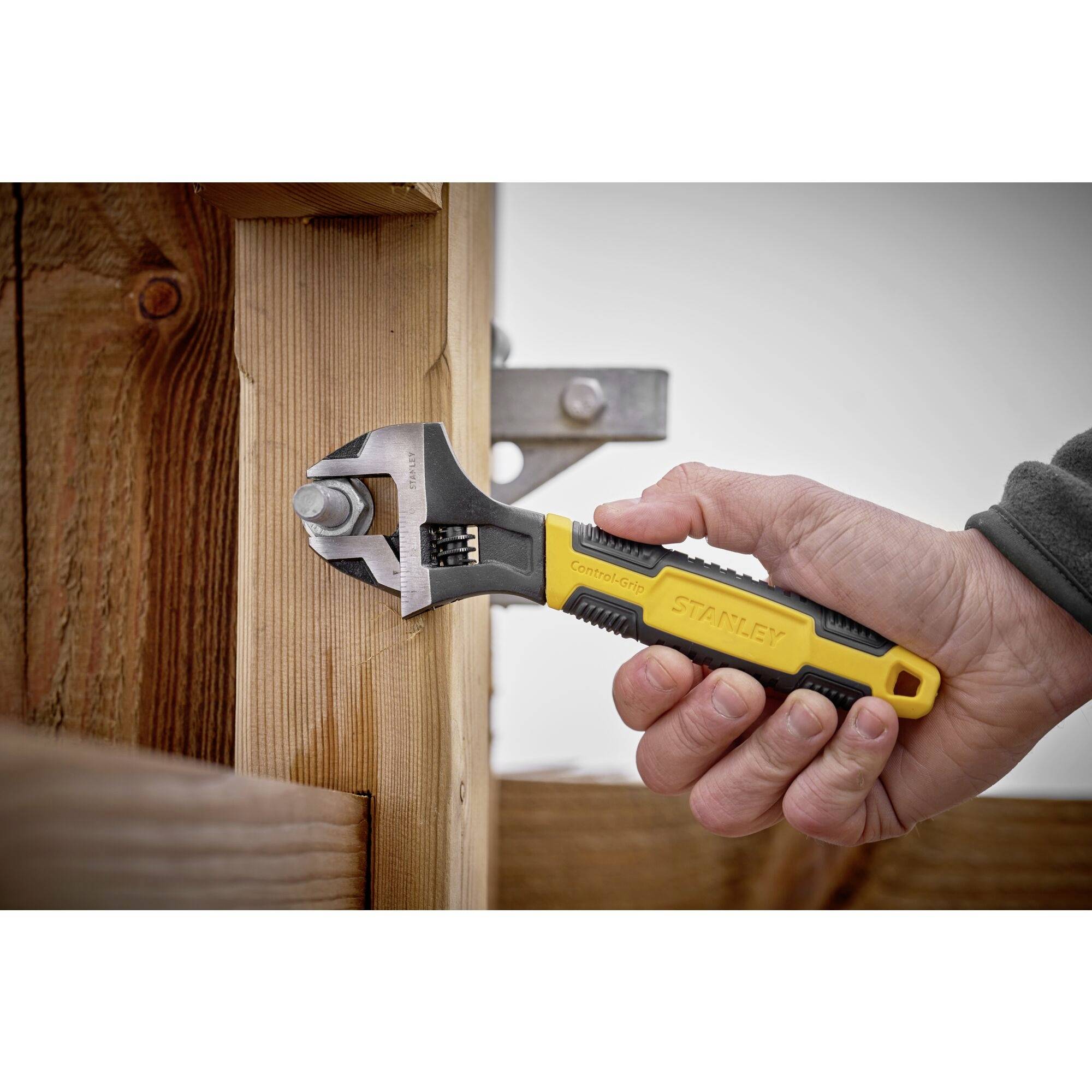 A hand is holding an adjustable spanner to tighten a screw in a wooden board. The spanner is coloured yellow and black.