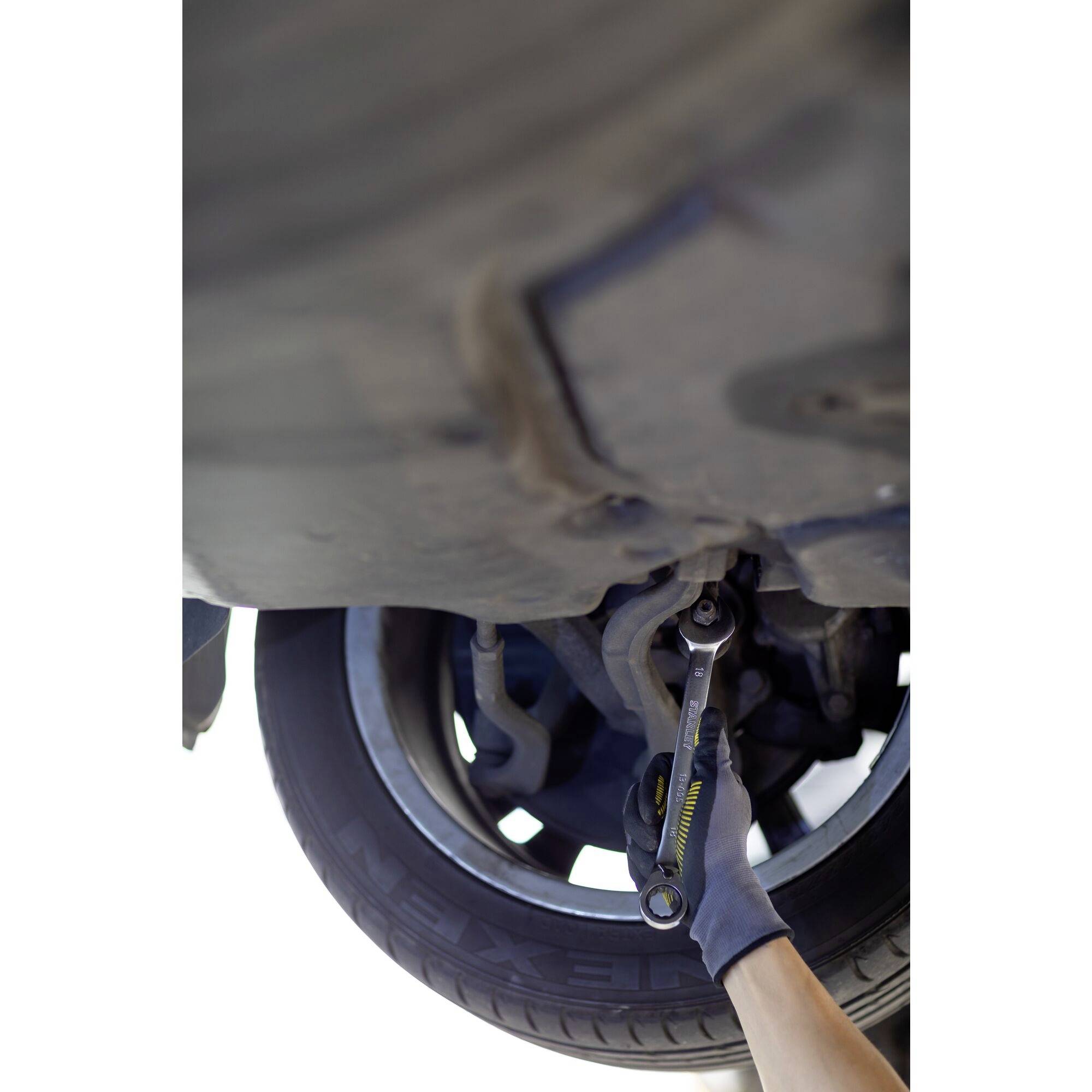A hand in a glove repairs the mechanics under a raised car with a spanner, with the wheel visible.