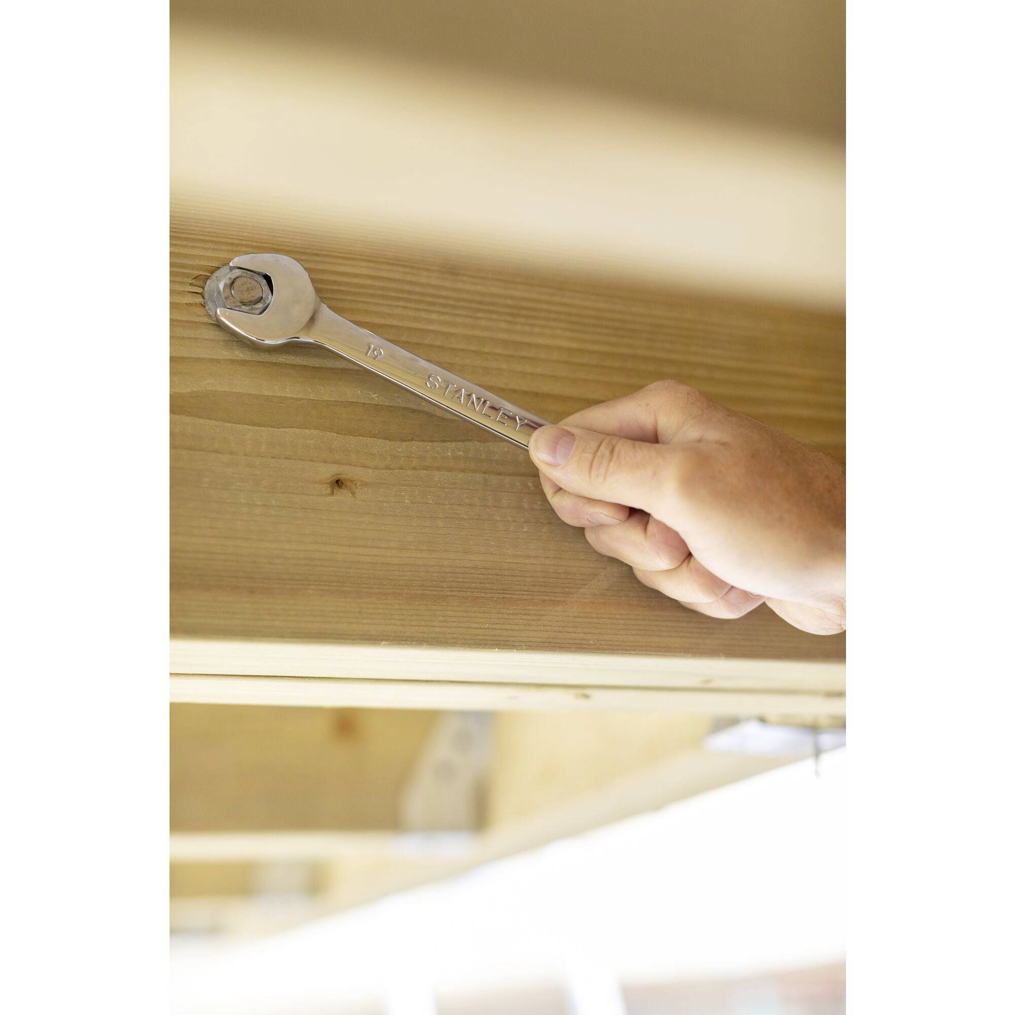 A hand holds a spanner pressed against a wooden surface to tighten a nut.