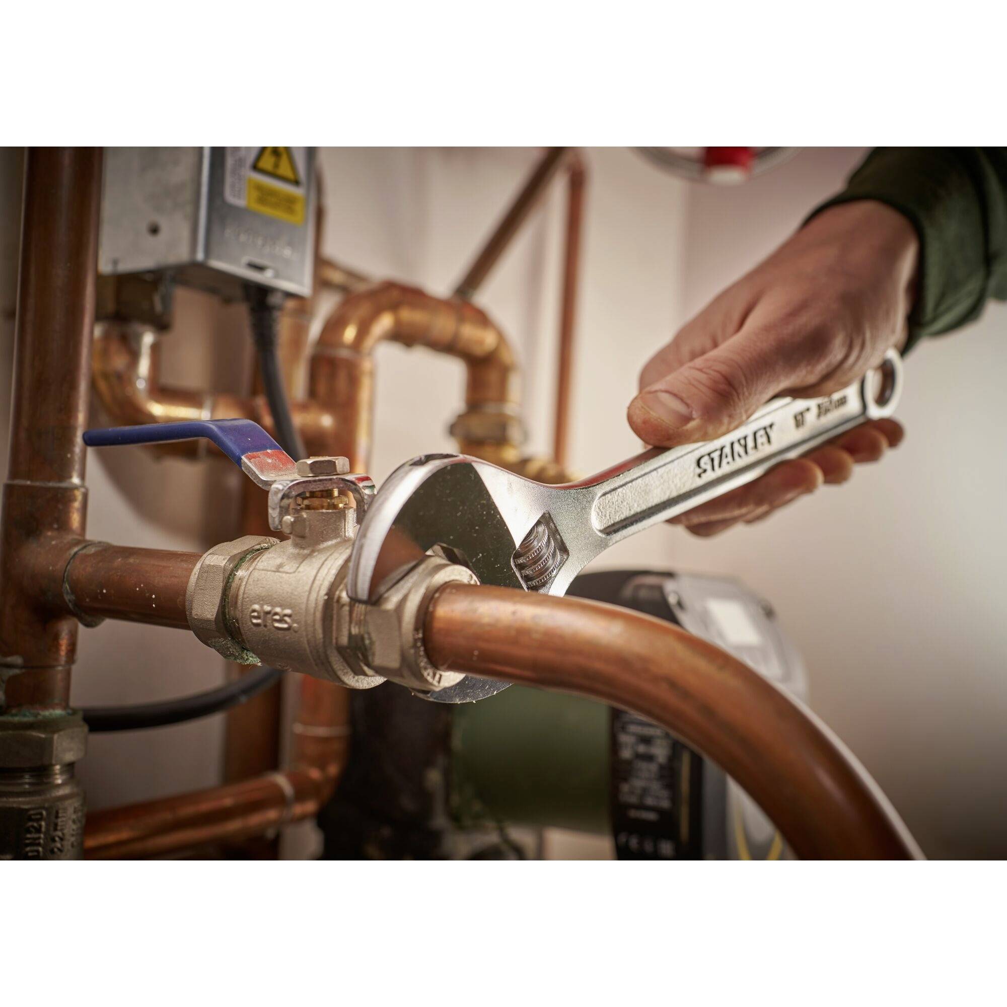 A hand is tightening a pipe fitting with a spanner. Copper pipes are visible.