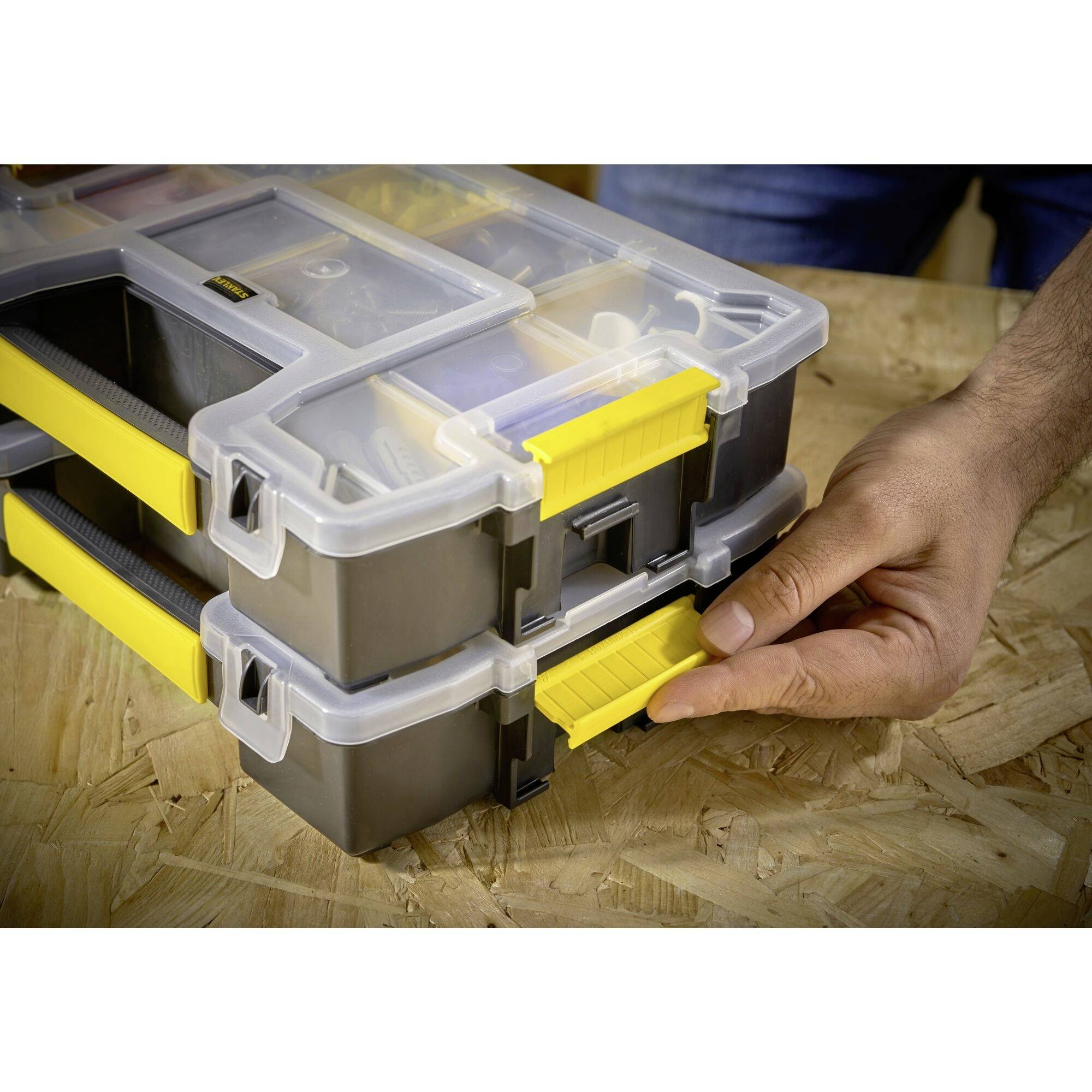 A hand opens a plastic toolbox with yellow clasps on a wooden table. The box has multiple compartments.