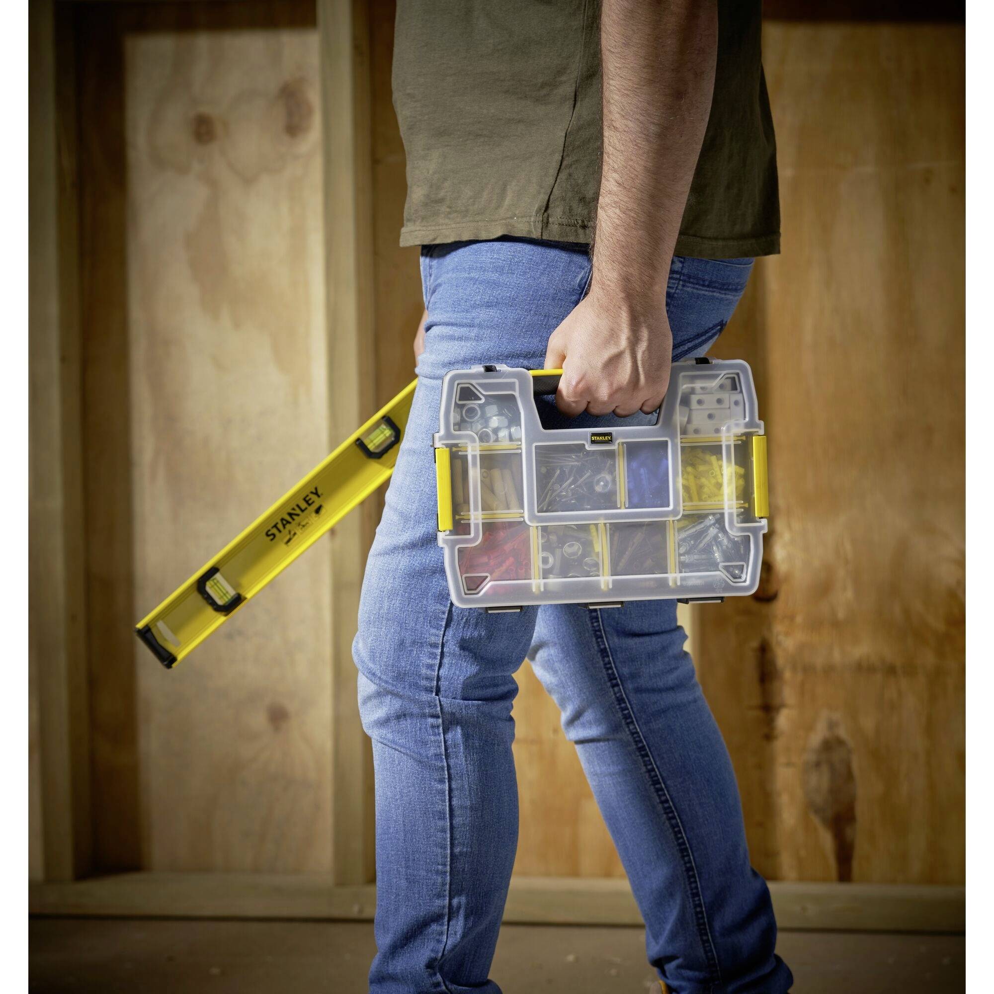 A person is carrying a spirit level and a toolbox with various tools. Wooden walls are visible in the background, suggesting a construction site.