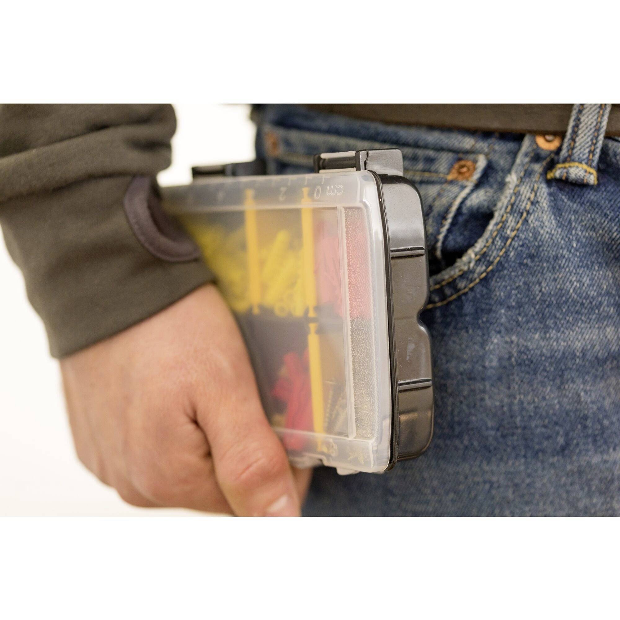 A person is holding a transparent box with small yellow and red objects near their trouser pocket.