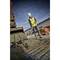A construction worker in protective clothing is spreading concrete with a tool next to a digger on a building site with stacks of bricks in the background.