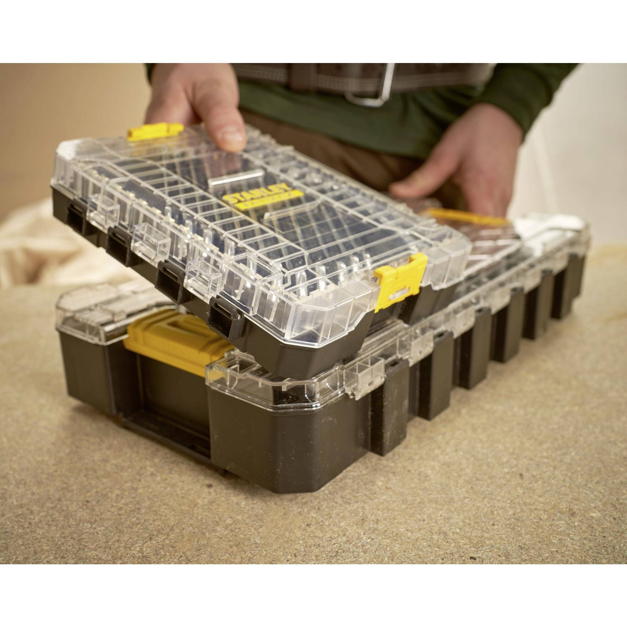A person lifts a transparent storage box with yellow closures from a table, which is suitable for tools or small parts.