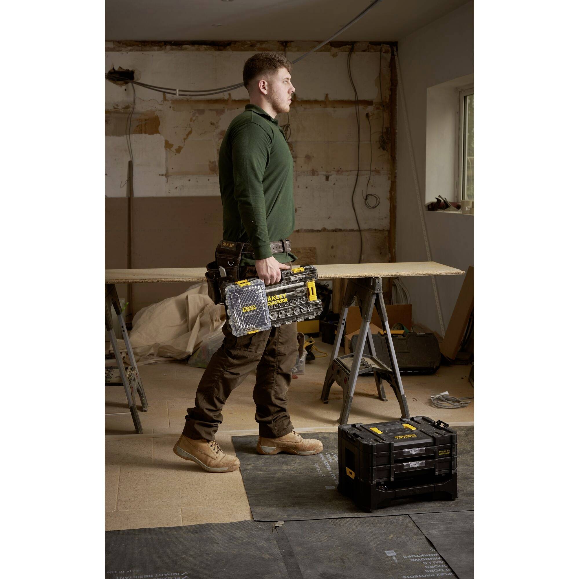 A man in workwear is standing in a workshop, holding a tool case in his hand. Tools and materials are visible in the background.