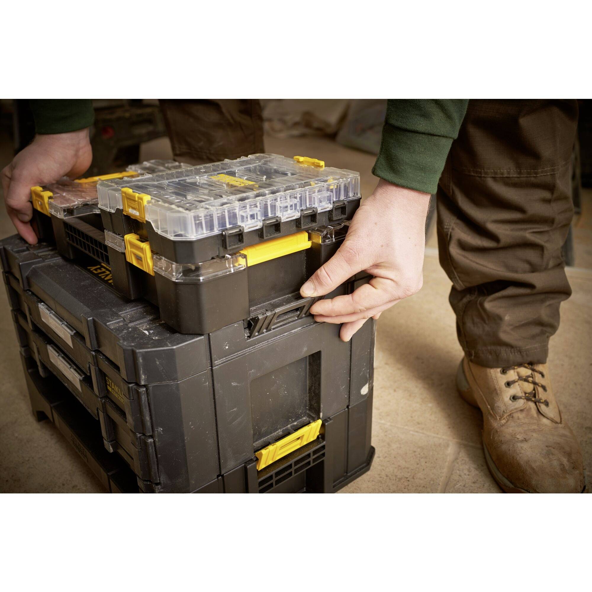 A person is stacking robust, black tool boxes with yellow handles. Their hands are connecting one box to another.