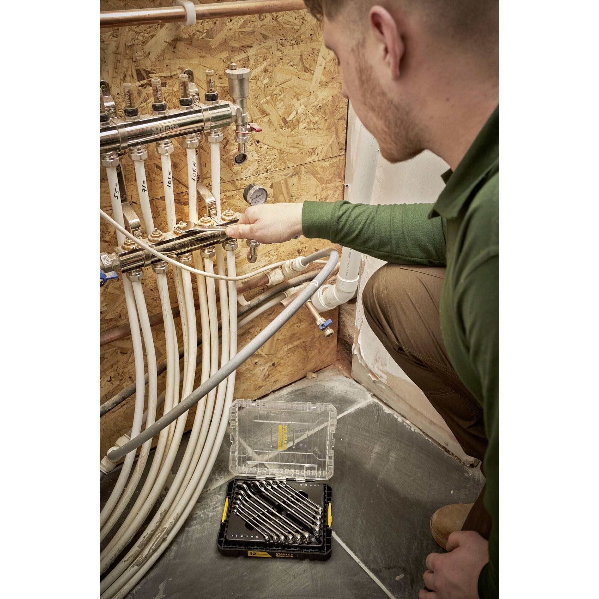 A man is working on a distribution box with several pipes in the boiler room. An open toolset lies beside him on the floor.