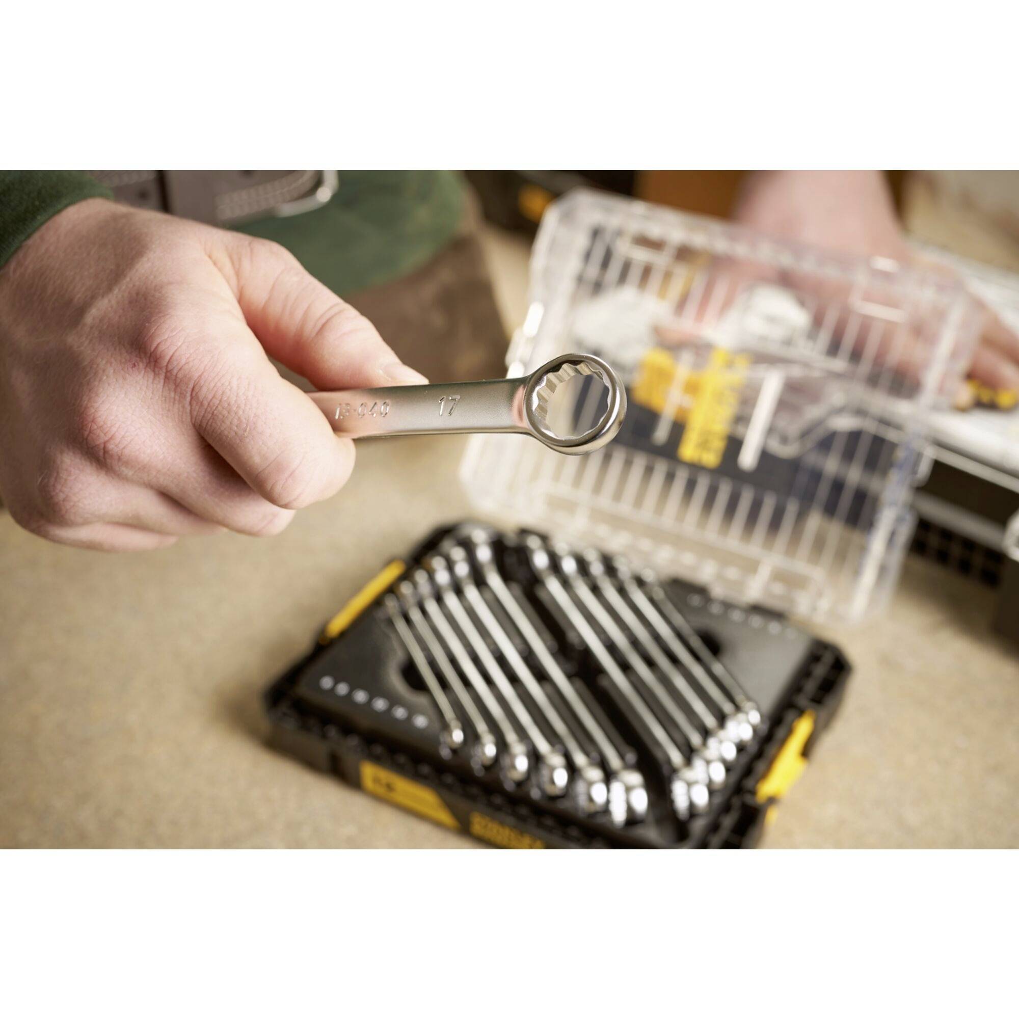 A person is holding a ring spanner over an opened set of spanners. The background shows various tools and a toolbox.